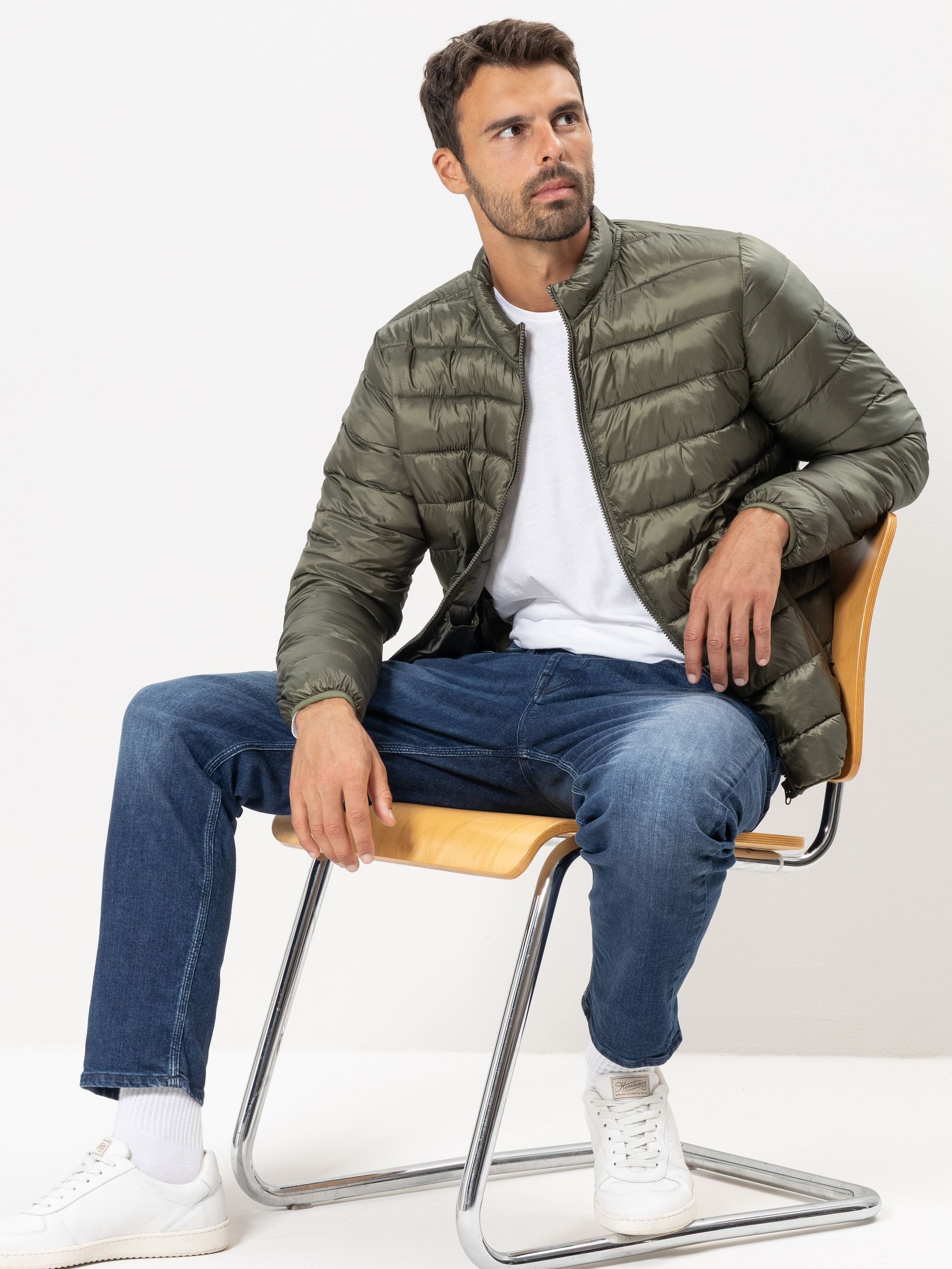 Ein Mann, der eine grüne Pufferjacke, ein weißes T-Shirt, blaue Jeans und weiße Turnschuhe trägt, sitzt auf einem hellbraunen Stuhl vor einem schlichten weißen Hintergrund. Er blickt mit entspanntem Gesichtsausdruck leicht zur Seite.