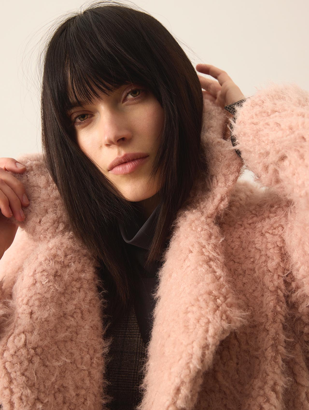 Eine Person mit glattem, schulterlangem dunklem Haar und Pony, die einen weichen, strukturierten hellrosa Mantel trägt, schaut direkt in die Kamera, während sie mit beiden Händen sanft den Kragen berührt.