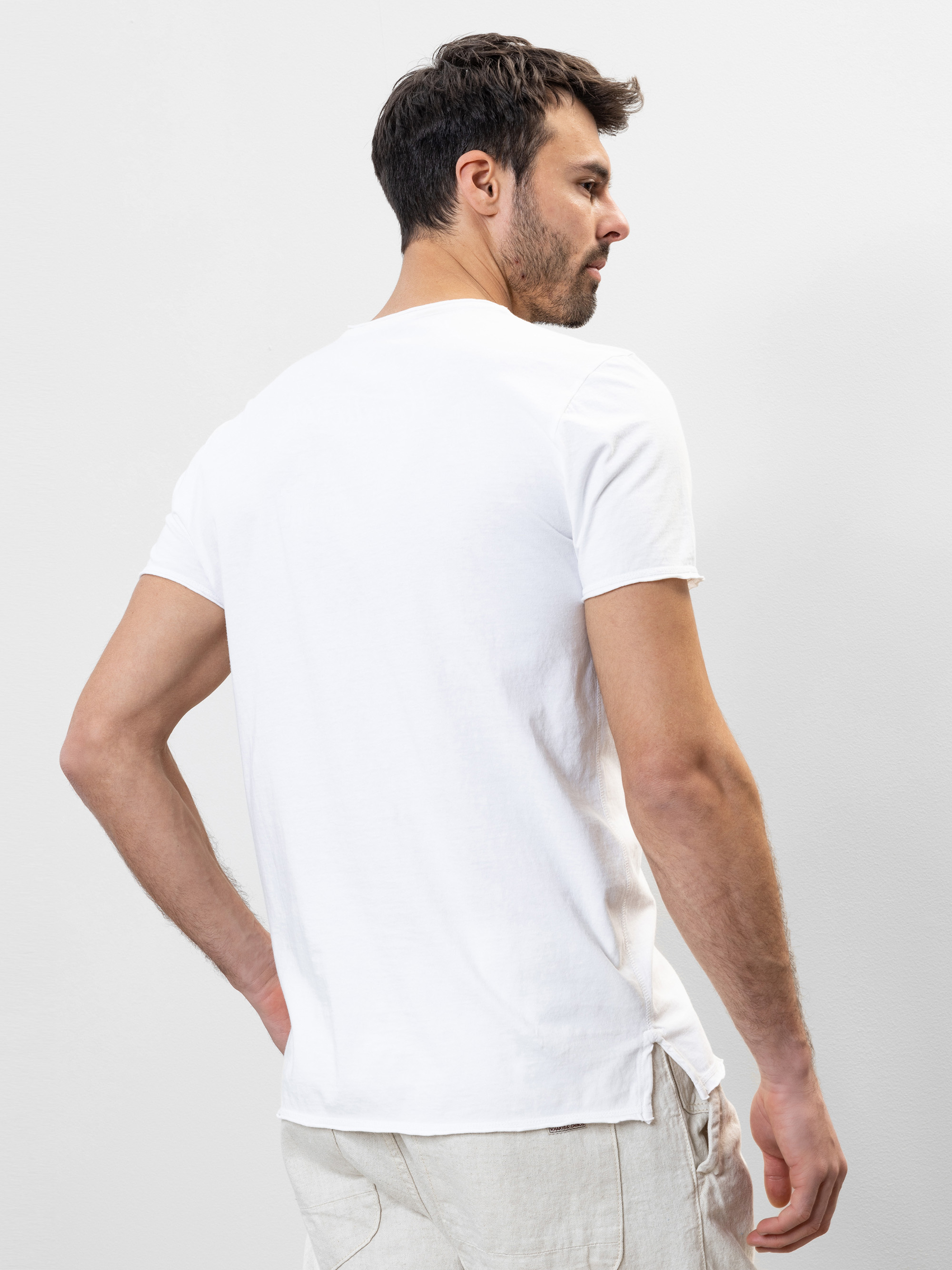 Ein Mann steht von der Kamera abgewandt und trägt ein schlichtes weißes, kurzärmeliges T-Shirt und eine helle Hose vor einem hellgrauen Hintergrund.