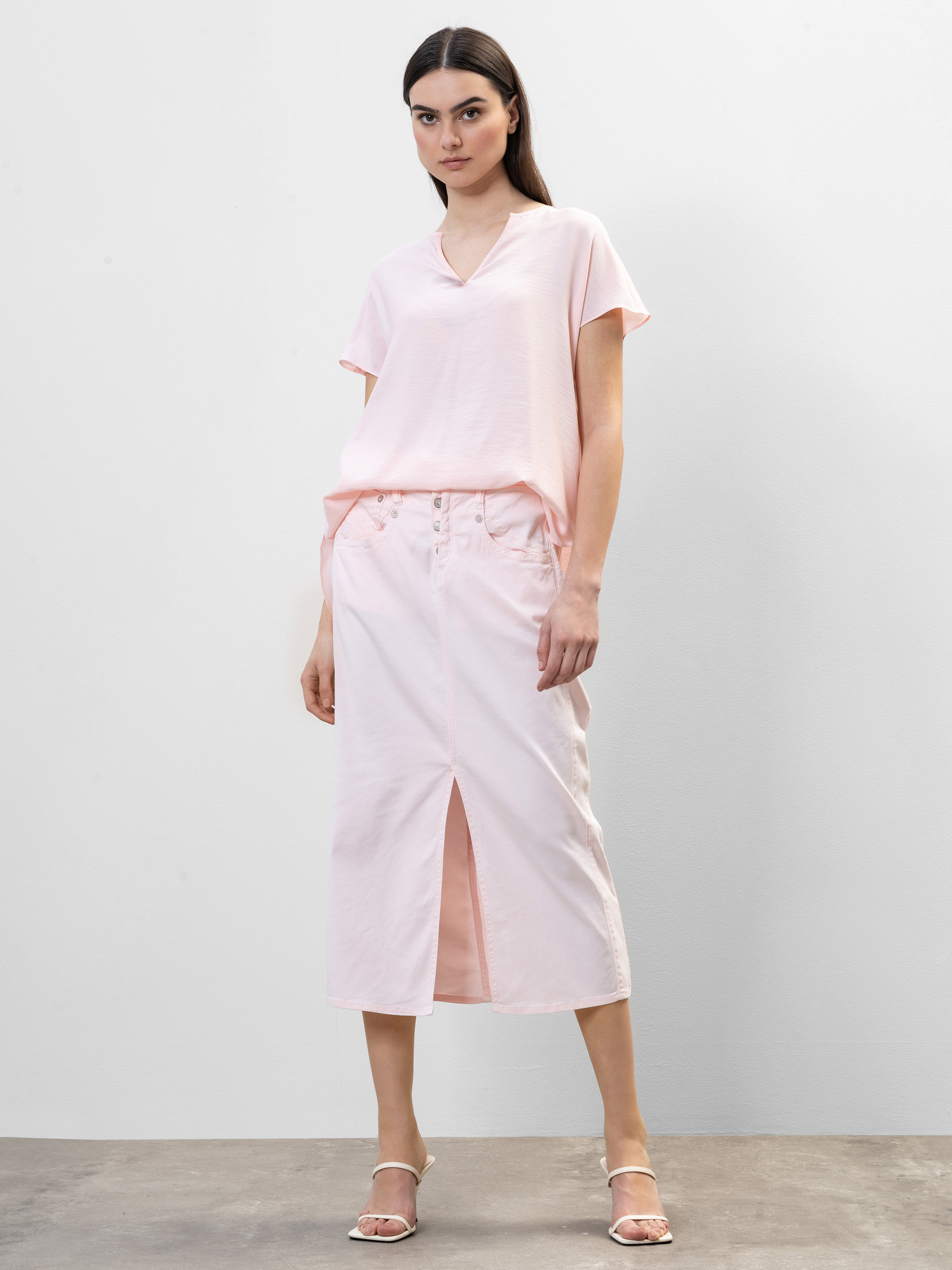 Eine Frau steht vor einem unifarbenen Hintergrund und trägt ein hellrosa, kurzärmeliges Oberteil und einen dazu passenden Midirock mit einem Schlitz an der Vorderseite, gepaart mit weißen, hochhackigen Sandalen.