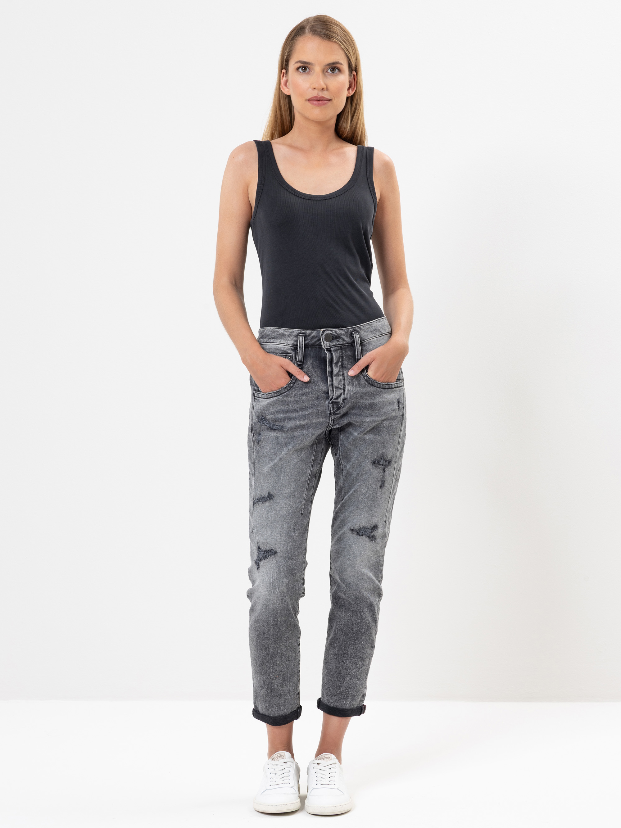 Eine Frau steht vor einem schlichten weißen Hintergrund und trägt ein ärmelloses schwarzes Oberteil, graue Jeans mit aufgerollten Bündchen und weiße Turnschuhe. Sie hat die Hände in den Taschen und blickt direkt in die Kamera.