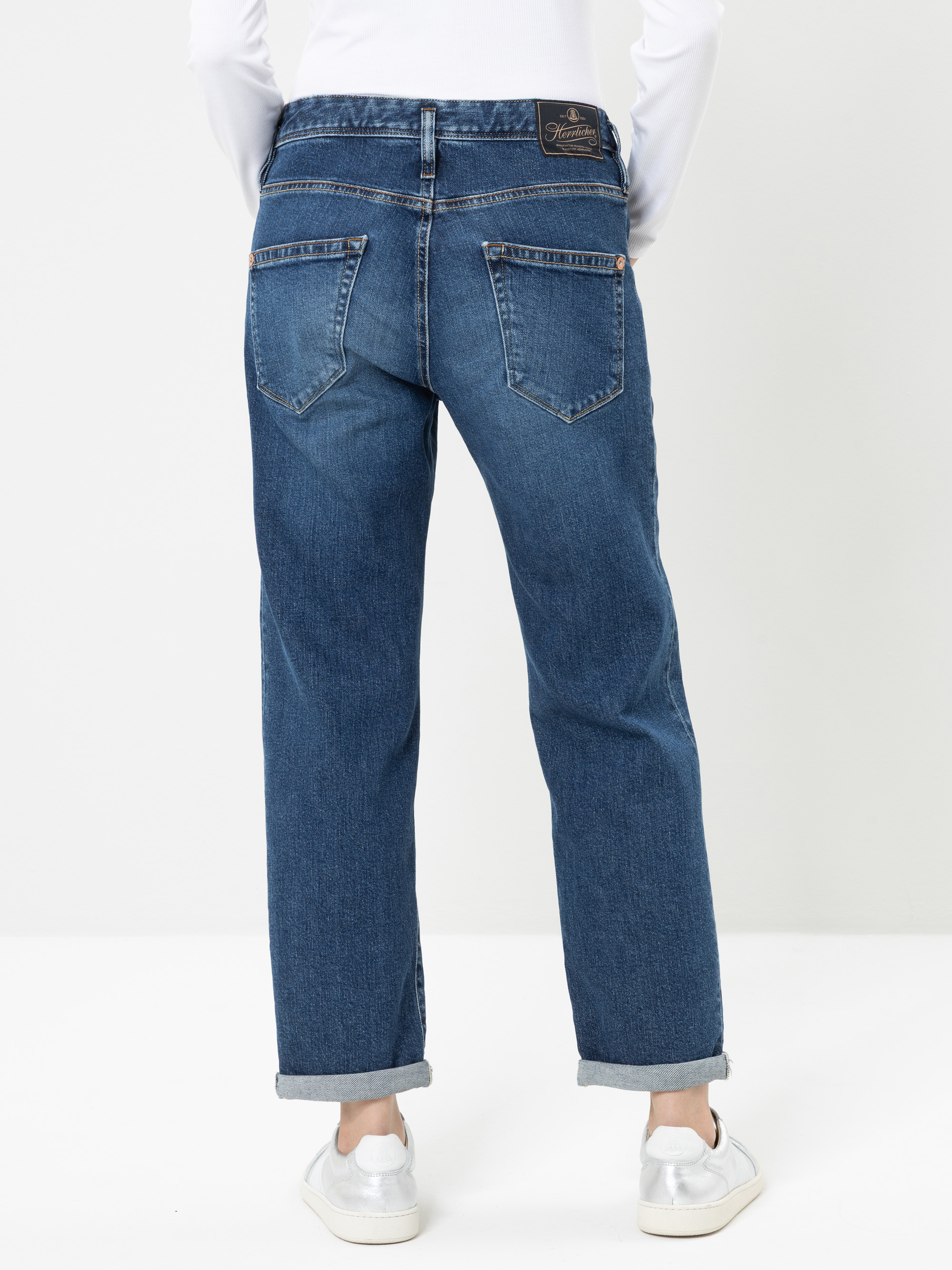 Eine Person, die eine blaue Jeans mit geradem Bein und umgeschlagenen Säumen, ein weißes Langarmhemd und weiße Turnschuhe trägt, steht vor einem schlichten, hellen Hintergrund und wird von hinten fotografiert.