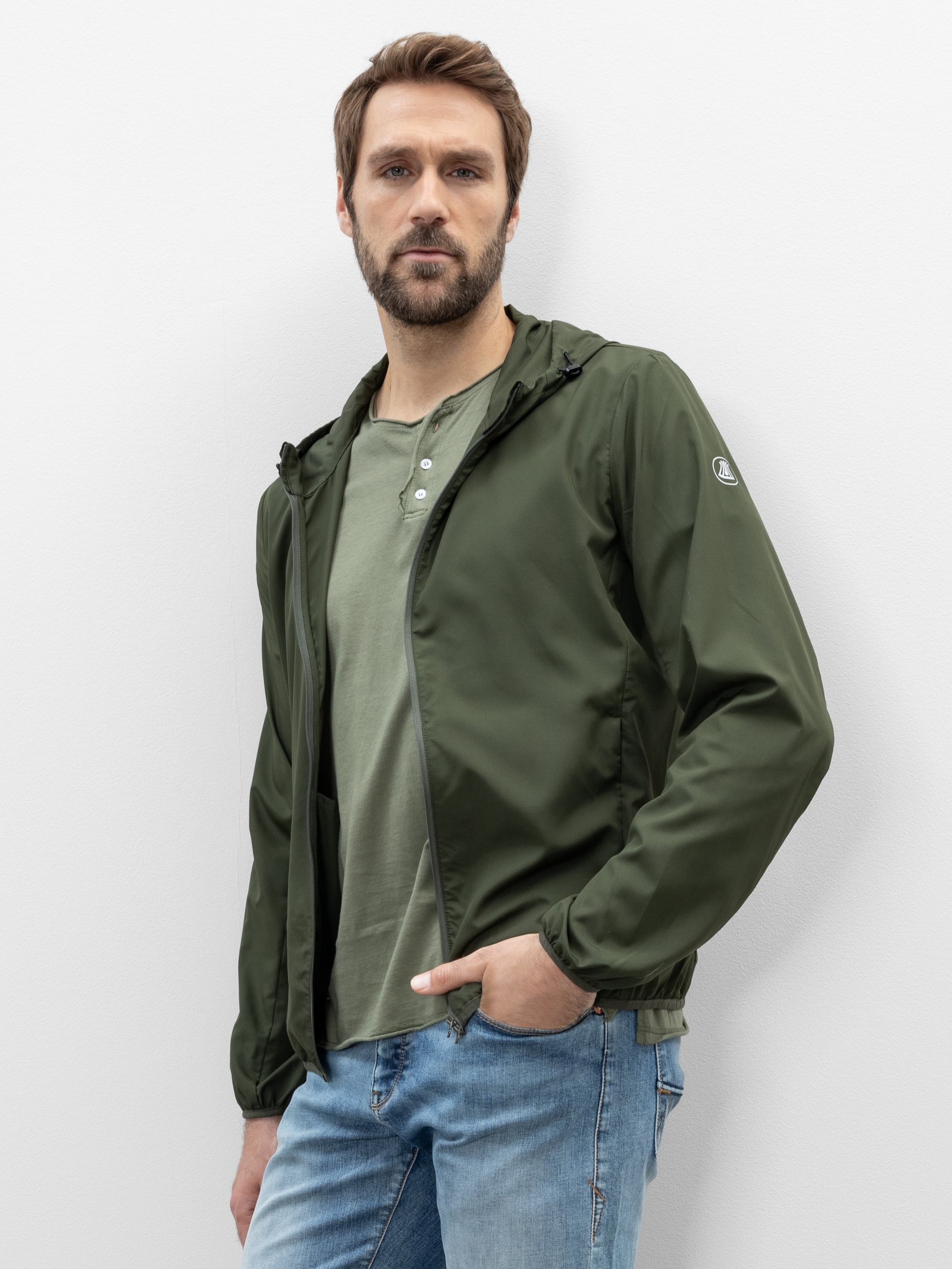 Ein Mann mit hellbraunem Haar und einem Bart trägt eine grüne Jacke über einem hellgrünen Hemd und blaue Jeans. Er steht vor einem schlichten weißen Hintergrund, hat eine Hand in der Tasche und schaut in die Kamera.