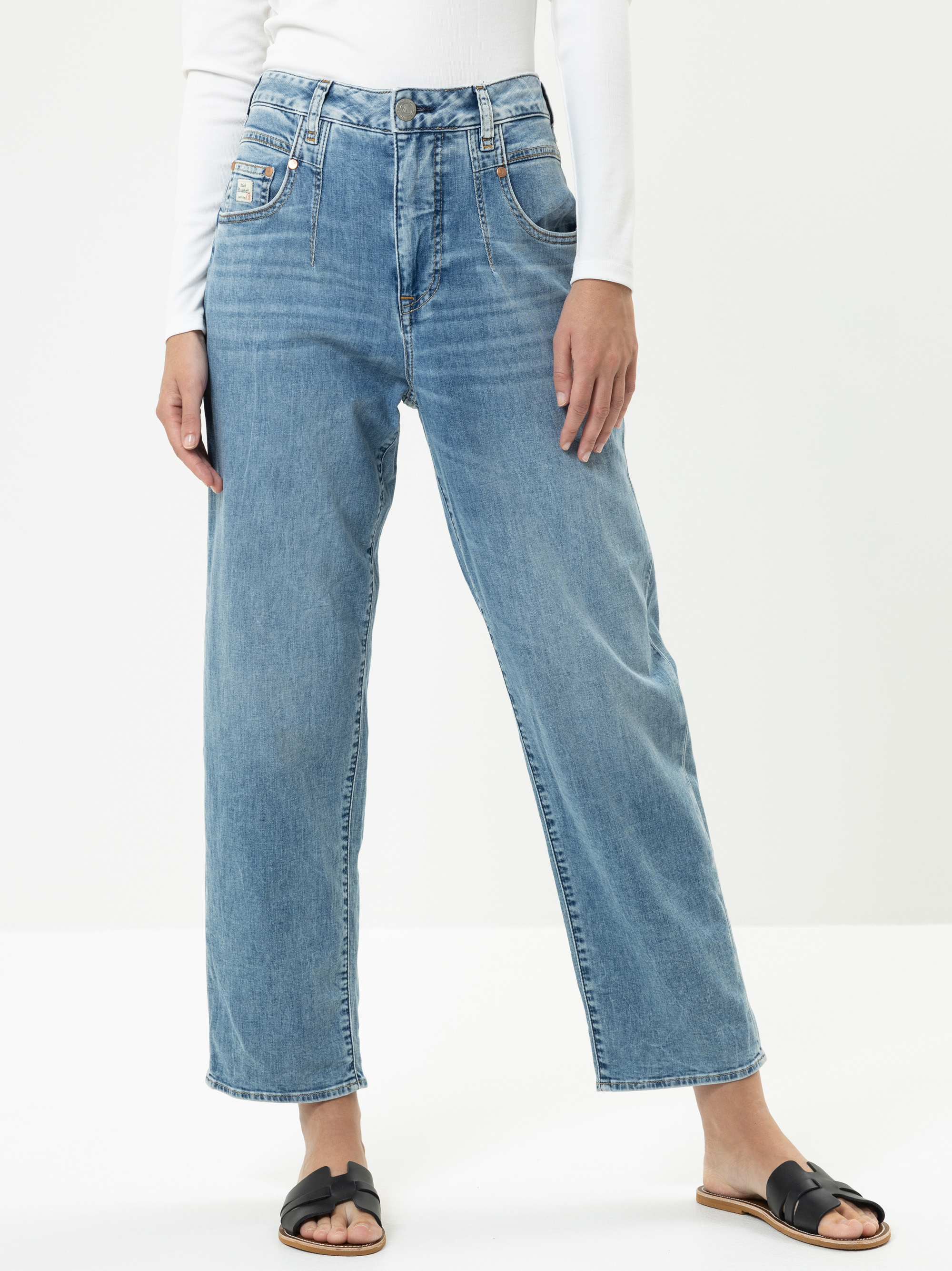 Person, die hochtaillierte, locker sitzende Bluejeans, ein weißes langärmeliges Oberteil und schwarze Slide-Sandalen trägt und vor einem schlichten weißen Hintergrund steht. Das Gesicht der Person ist nicht sichtbar.