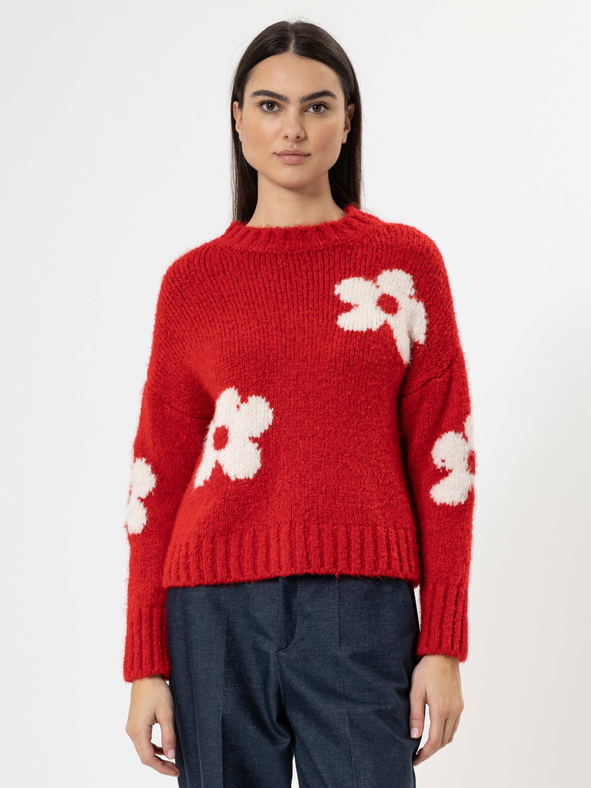 Eine Frau mit glattem dunklem Haar trägt einen roten Strickpulli mit großen weißen Blumenmustern und eine dunkle Hose, die vor einem schlichten weißen Hintergrund steht.