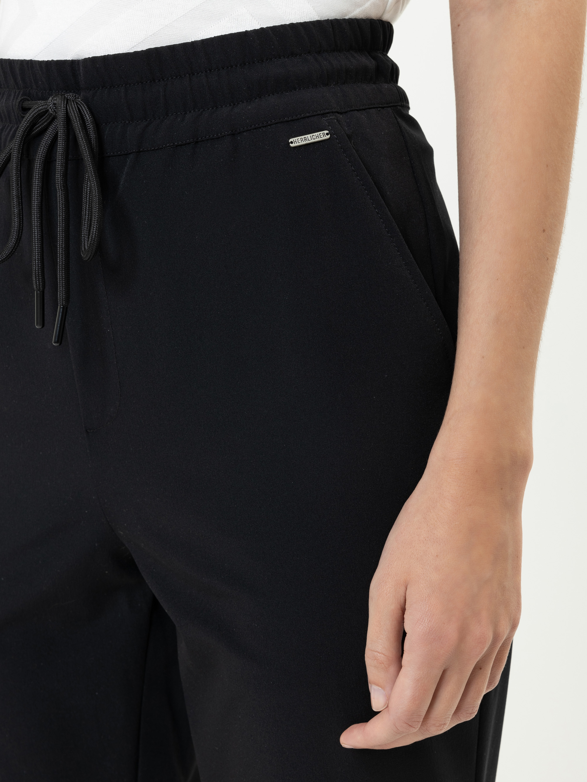 Eine Person, die eine schwarze Hose mit Kordelzug und ein weißes Hemd trägt, wobei eine Hand in der Nähe der Tasche ruht. Die Hose hat ein kleines metallisches Markenzeichen in der Nähe des Hosenbundes.