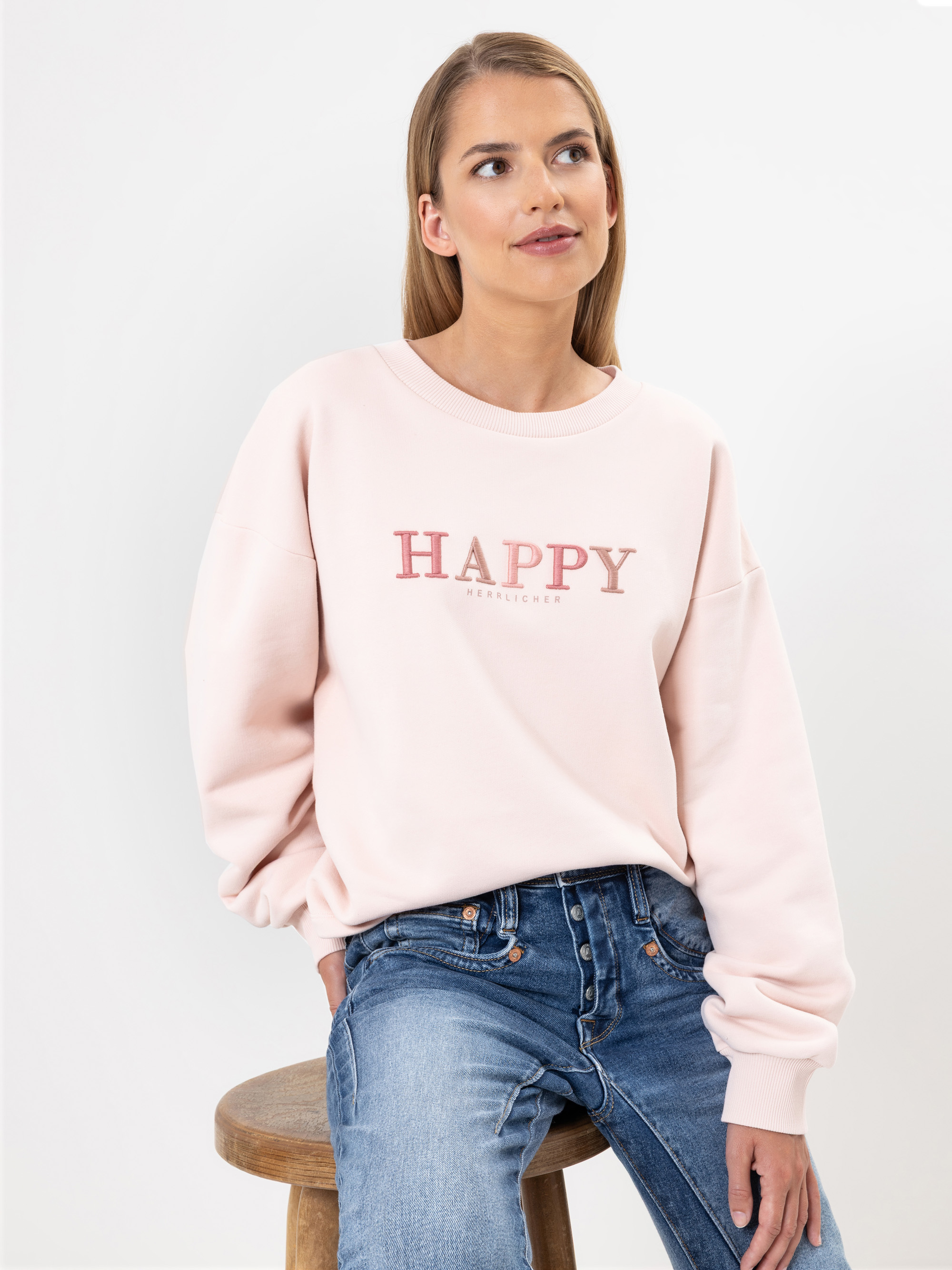 Eine Frau mit glattem, hellbraunem Haar sitzt auf einem Holzschemel, trägt ein hellrosa Sweatshirt mit dem Wort HAPPY in fetten Buchstaben und eine blaue Jeans, schaut leicht nach oben und zur Seite und lächelt sanft.