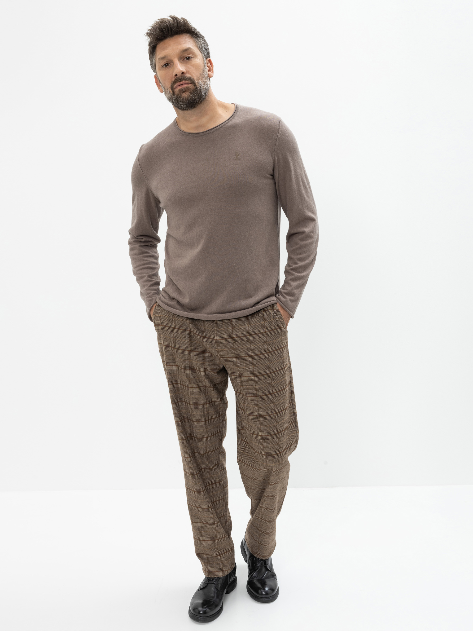 Ein Mann mit kurzen dunklen Haaren und einem Bart steht vor einem schlichten weißen Hintergrund. Er trägt ein taupefarbenes langärmeliges Hemd, eine braun karierte Hose und schwarze Schuhe, die Hände in den Taschen.