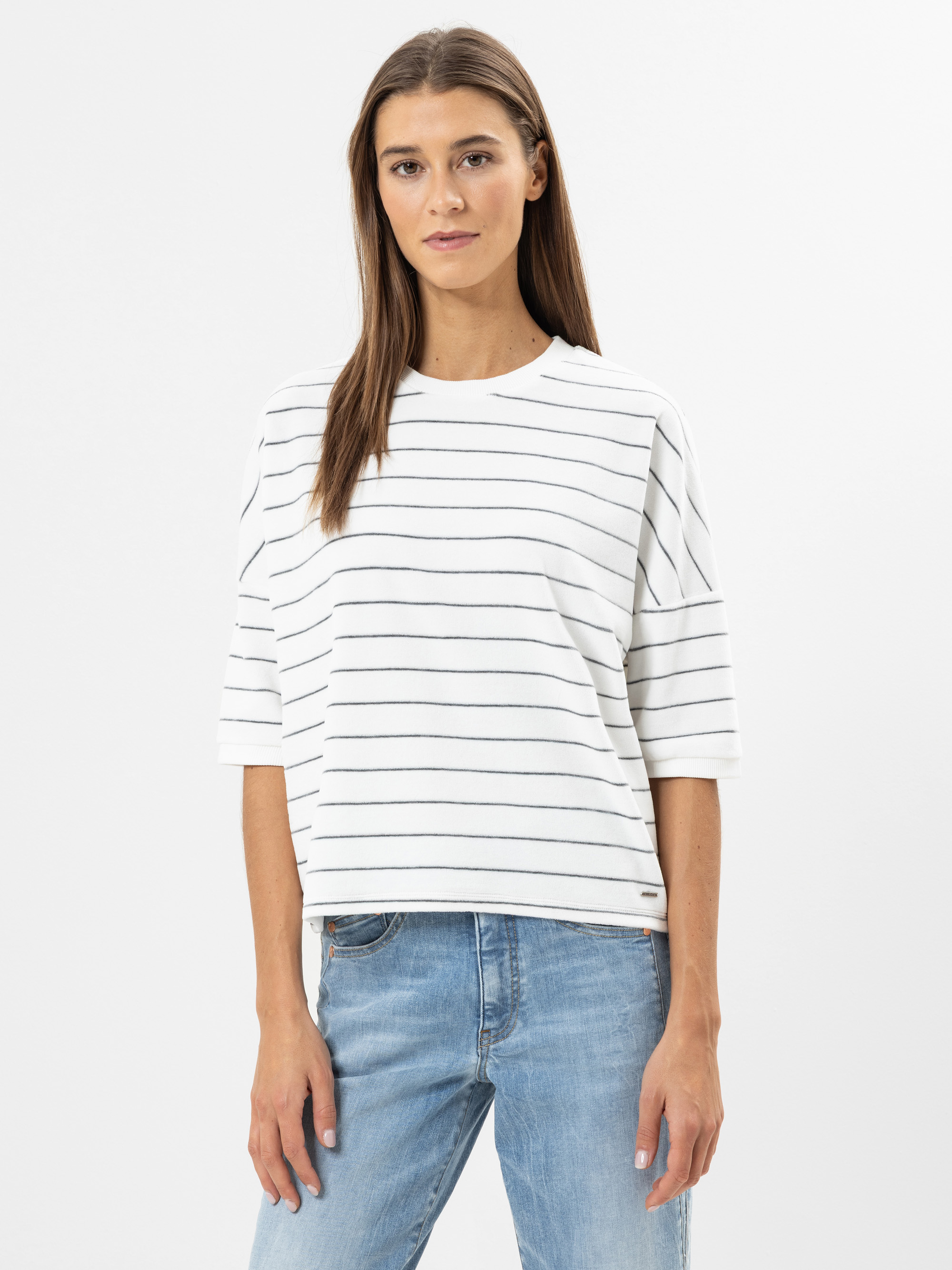 Eine Frau steht vor einem weißen Hintergrund und trägt ein weißes, kurzärmeliges, locker sitzendes Hemd mit dünnen schwarzen Querstreifen und hellblaue Jeans. Sie hat langes, glattes braunes Haar und einen neutralen Gesichtsausdruck.