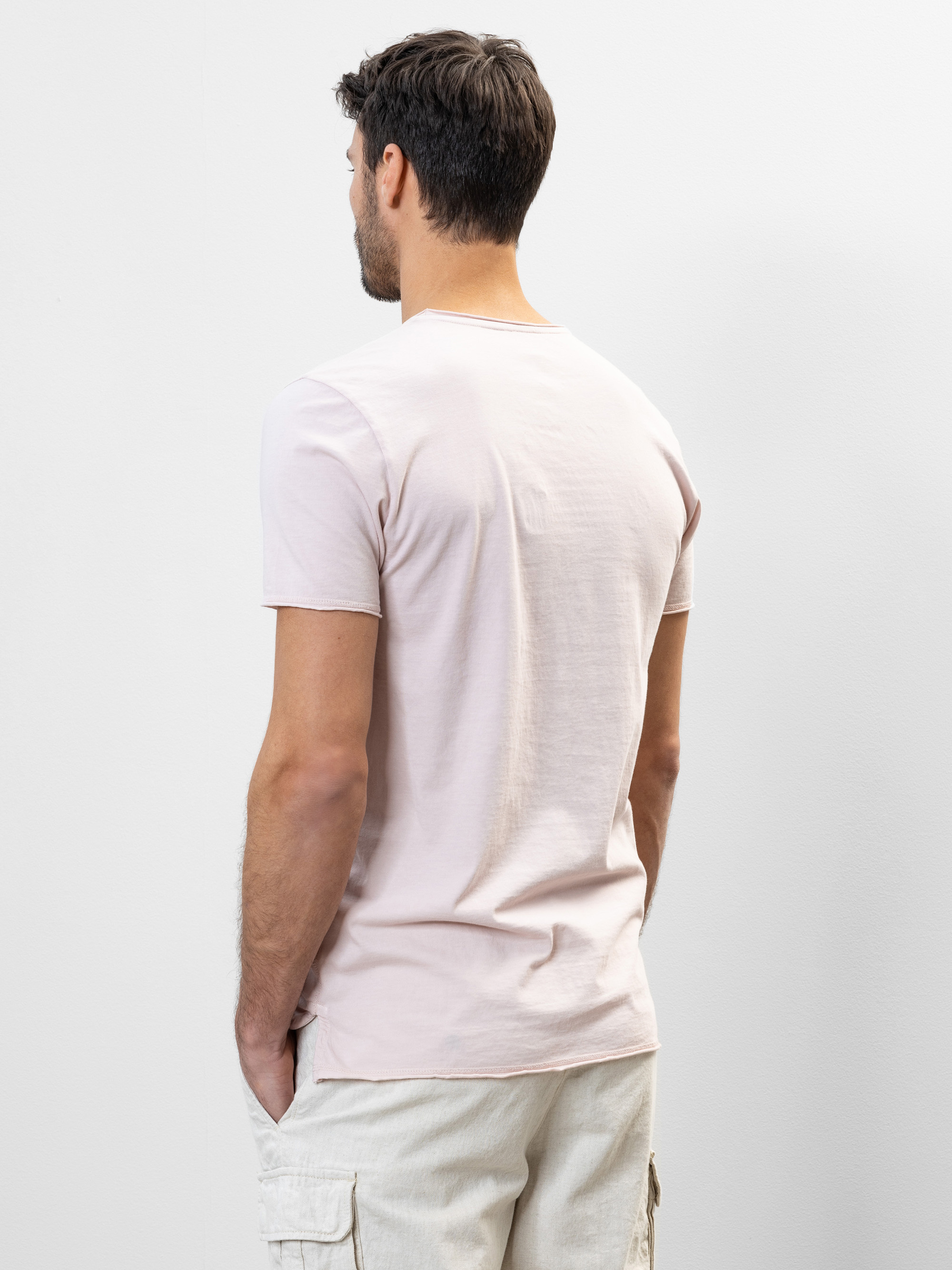 Ein Mann mit kurzen braunen Haaren steht mit dem Rücken zur Kamera, trägt ein hellrosa T-Shirt und beigefarbene Cargoshorts, die Hände in den Taschen, vor einem schlichten hellen Hintergrund.