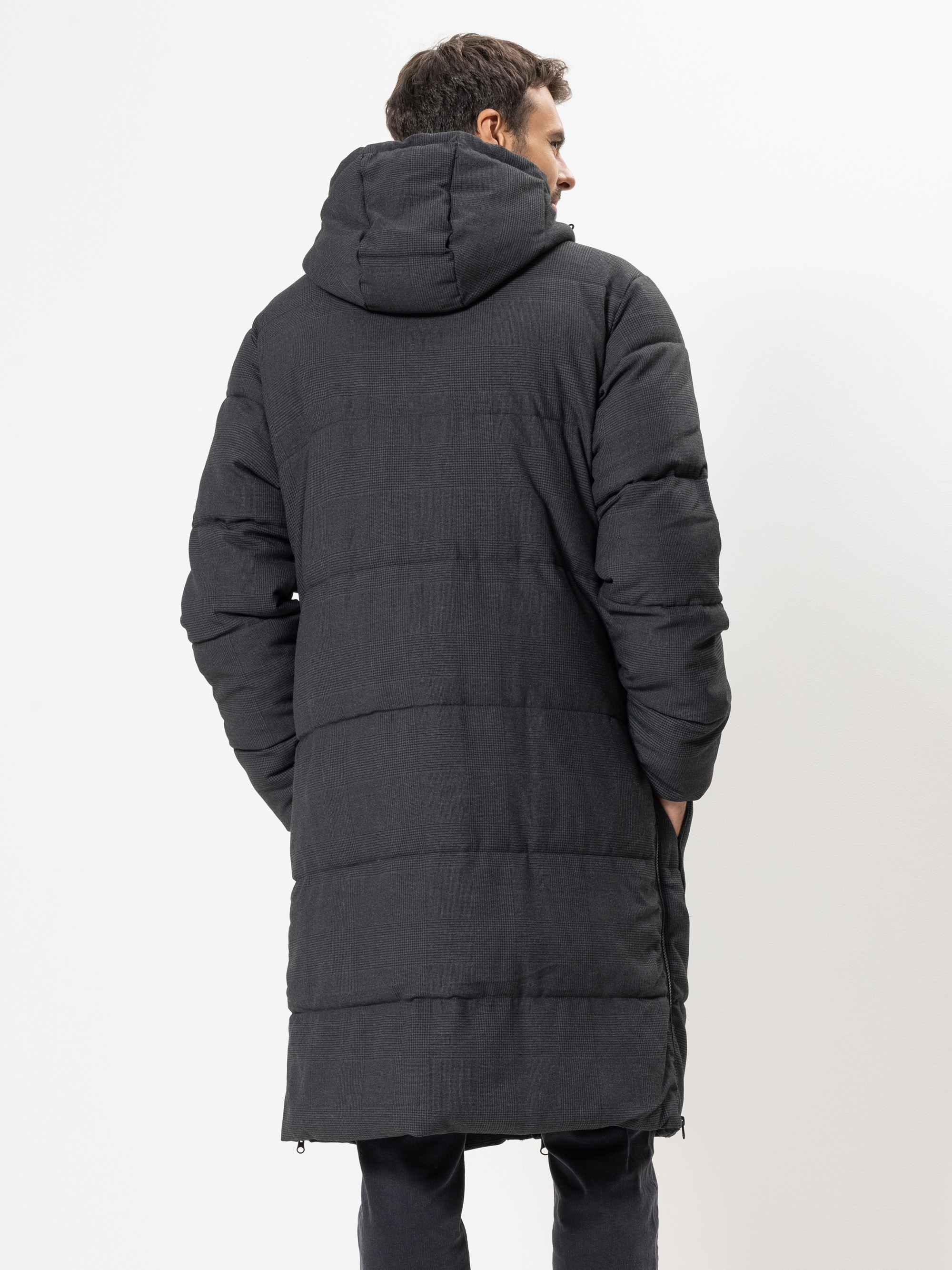 Ein Mann steht mit dem Rücken zur Kamera und trägt einen langen, schwarzen wattierten Wintermantel mit Kapuze. Der Mantel hat eine horizontale Steppung und reicht ihm bis zu den Knien. Der Hintergrund ist einfarbig weiß.