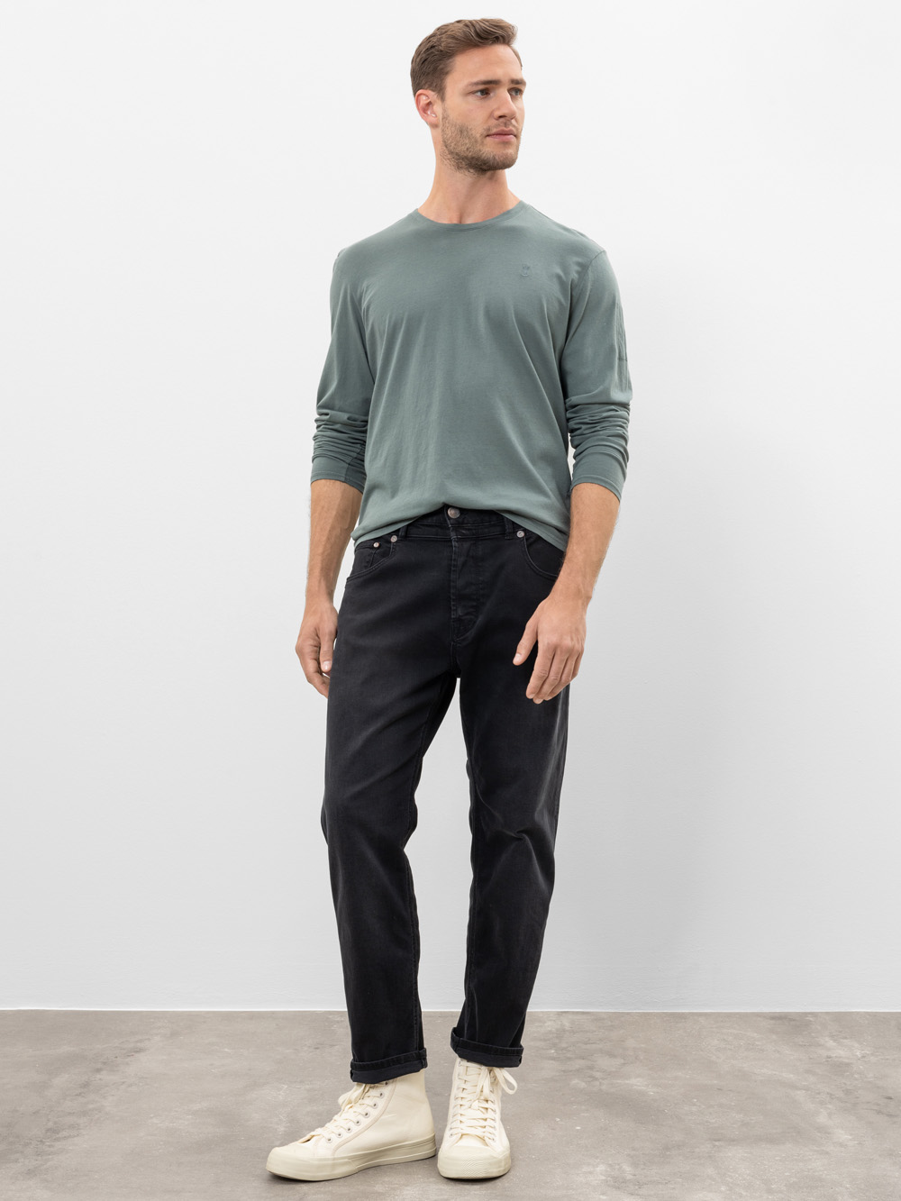 Ein Mann steht an einer schlichten weißen Wand. Er trägt ein hellgrünes, langärmeliges Hemd, schwarze Jeans und weiße Turnschuhe mit hohen Absätzen. Er blickt mit entspannter Haltung auf einem grauen Boden zur Seite.