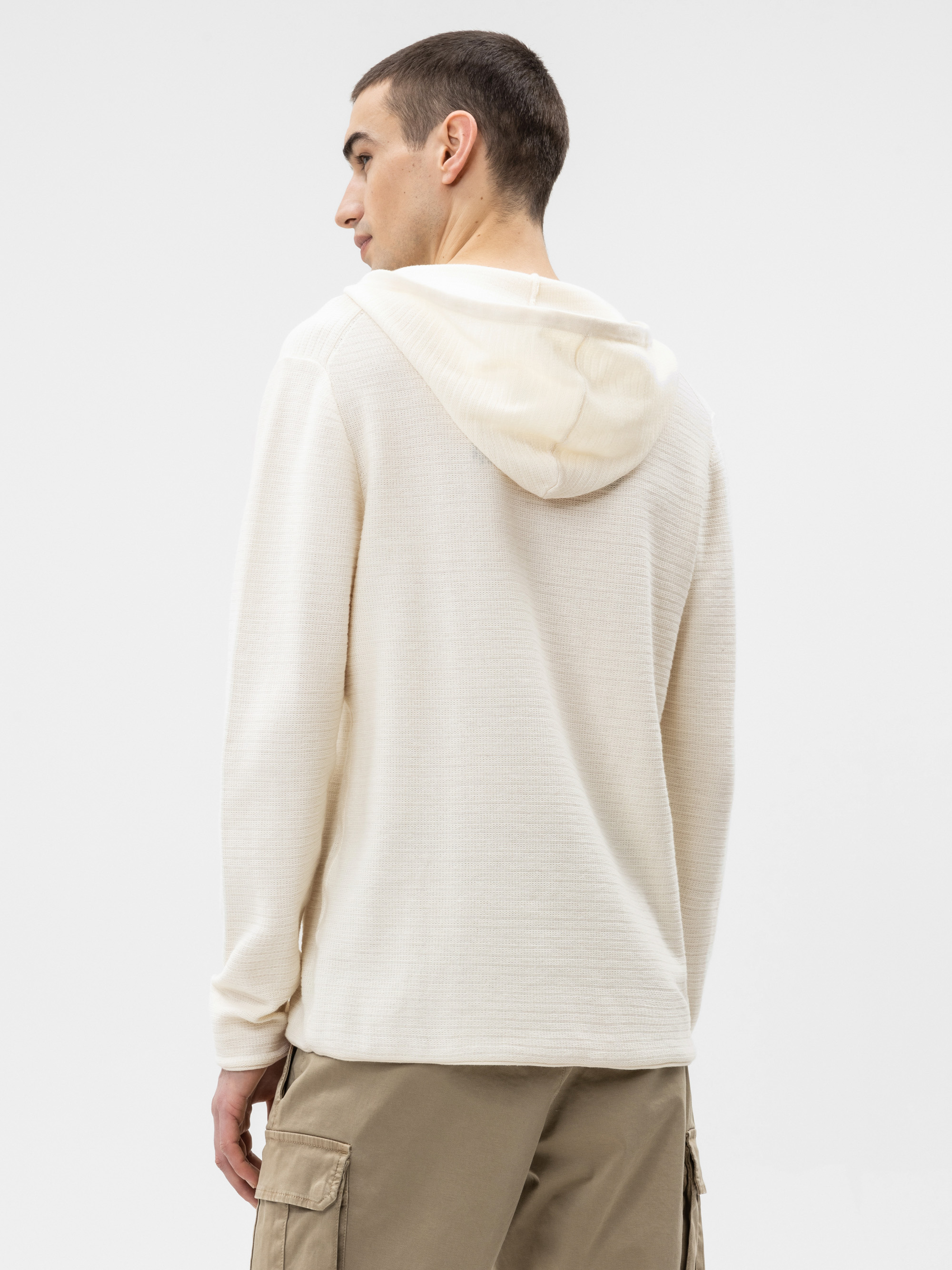 Ein Mann steht mit dem Gesicht zur Wand und trägt einen cremefarbenen, strukturierten Kapuzenpullover und eine hellbraune Cargohose vor einem schlichten weißen Hintergrund.