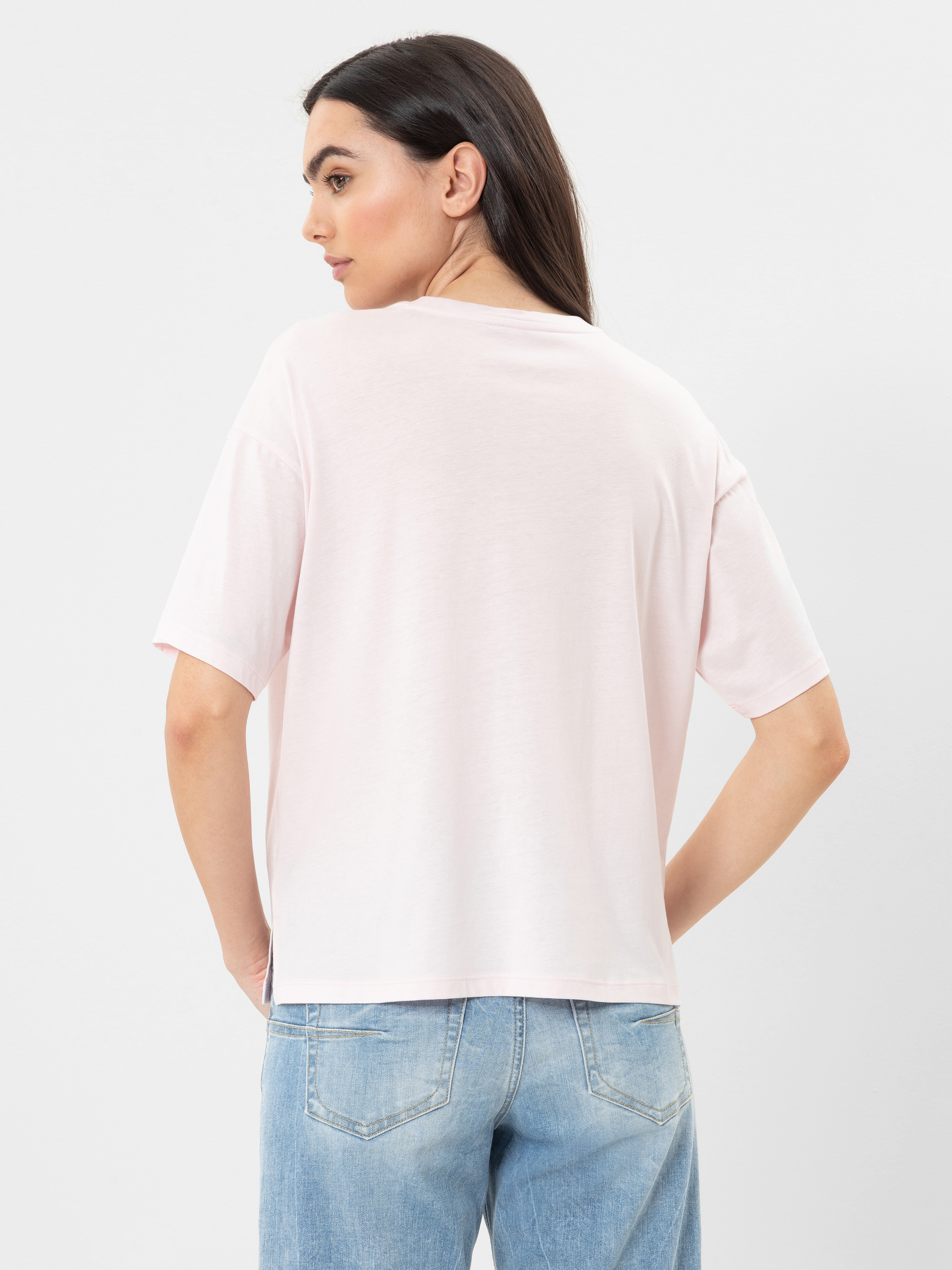 Eine Frau mit langen dunklen Haaren steht mit dem Rücken zur Kamera, trägt ein hellrosa kurzärmeliges T-Shirt und hellblaue Jeans vor einem schlichten weißen Hintergrund.