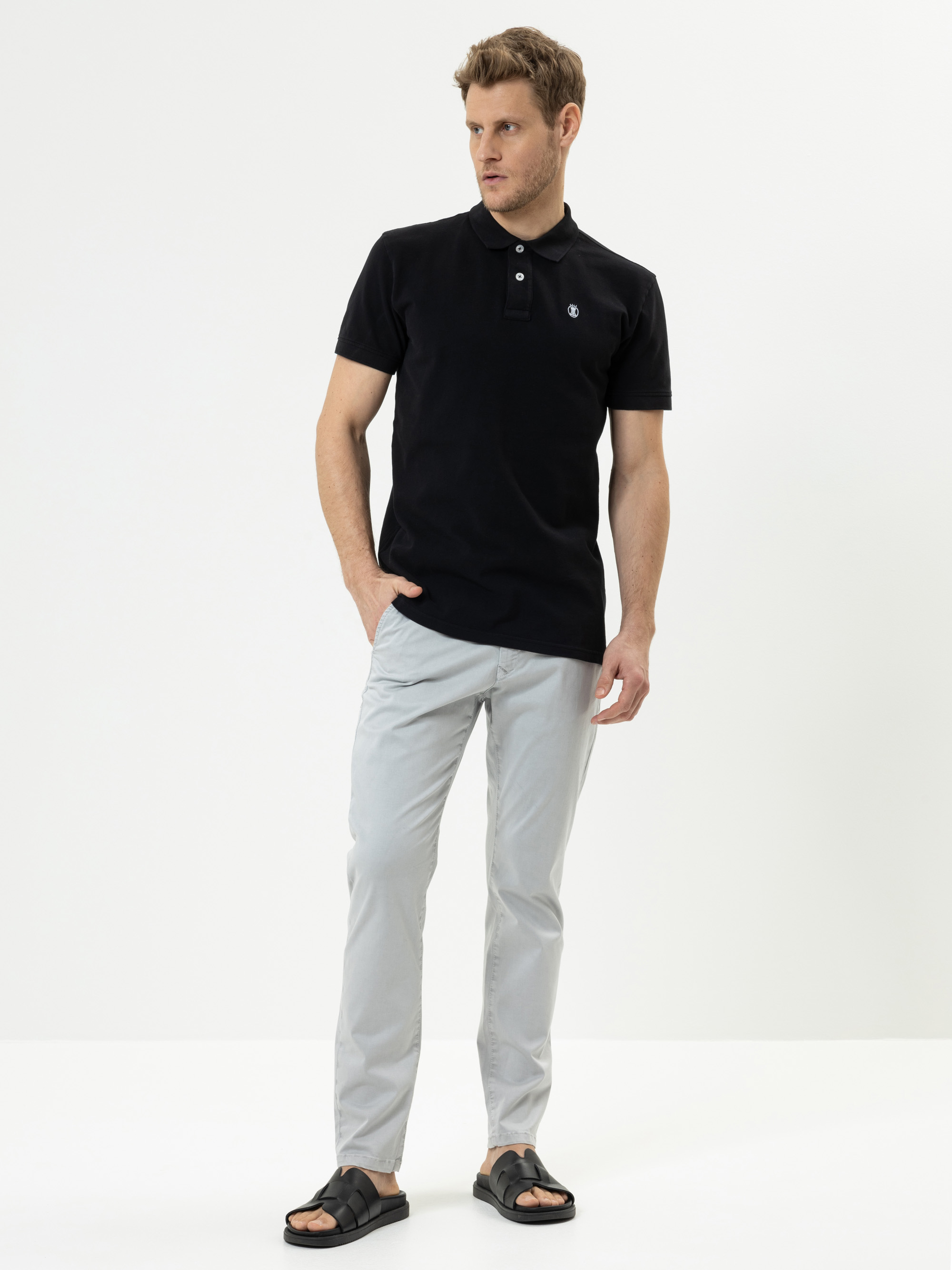 Ein Mann steht vor einem schlichten weißen Hintergrund und trägt ein schwarzes Poloshirt, eine hellgraue Hose und schwarze Sandalen. Er hat eine Hand in der Tasche und blickt zur Seite.