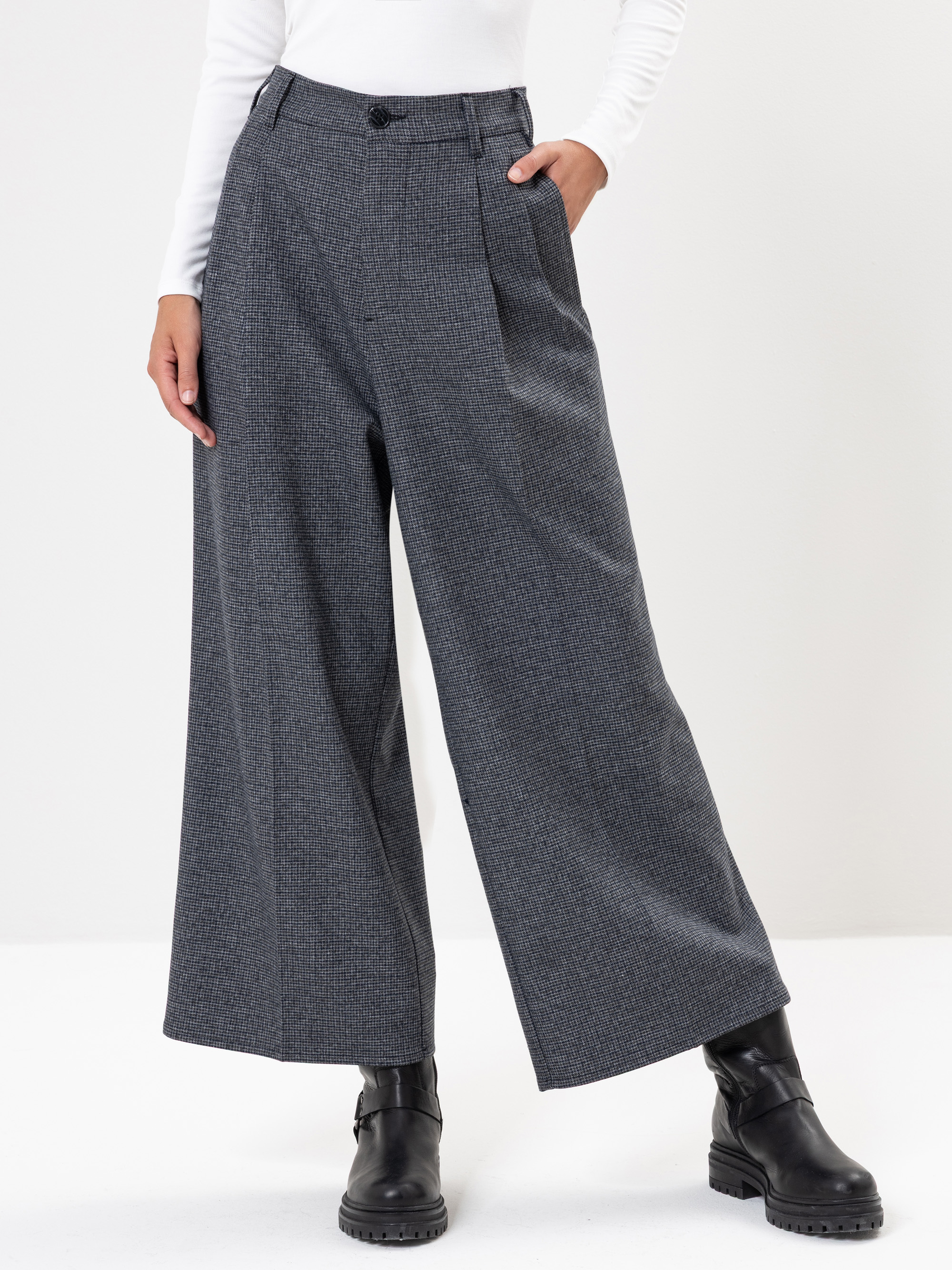 Eine Person, die eine weit geschnittene, grau karierte Hose, ein weißes, langärmeliges Oberteil und schwarze Stiefeletten trägt, steht vor einem schlichten weißen Hintergrund.