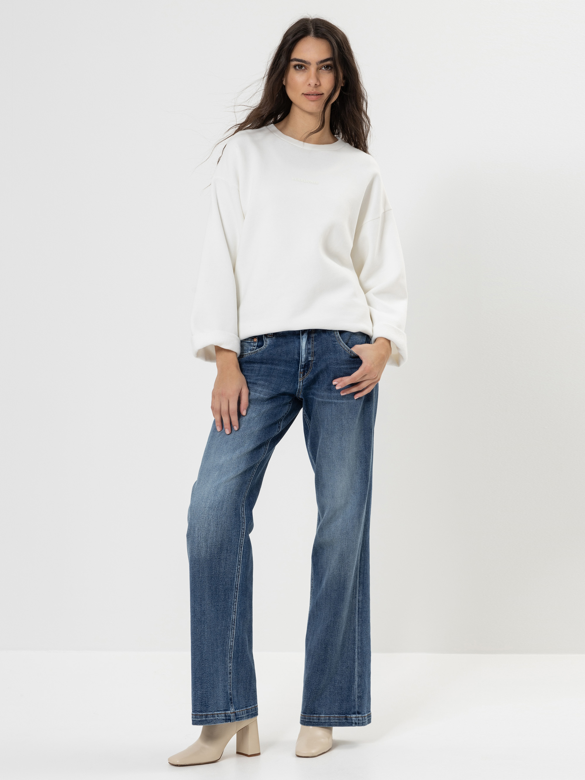 Eine Person mit langen dunklen Haaren trägt ein lockeres weißes Sweatshirt, blaue Jeans mit geradem Bein und beigefarbene Stiefel mit Absatz und steht vor einem schlichten weißen Hintergrund.