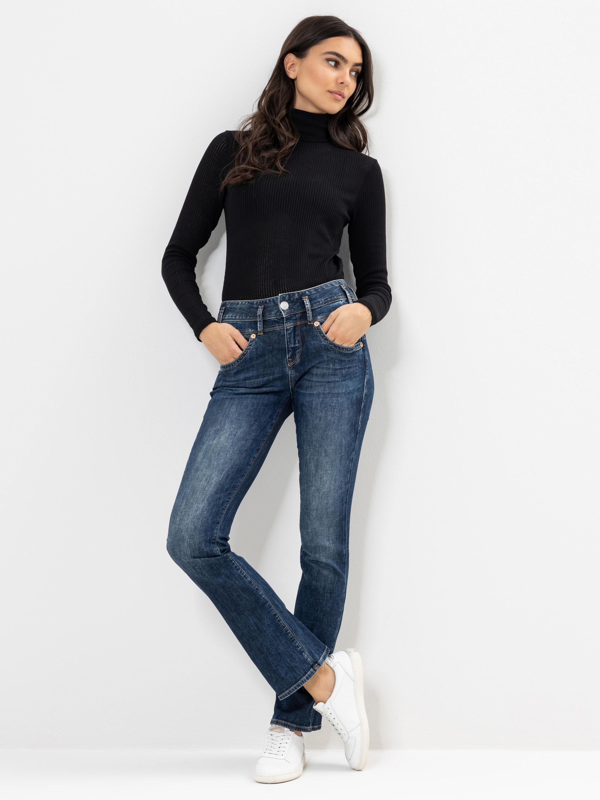 Eine Frau steht vor einem schlichten weißen Hintergrund und trägt einen schwarzen Rollkragenpullover, blaue Jeans und weiße Turnschuhe. Sie hat die Hände in den Hosentaschen und schaut mit entspanntem Gesichtsausdruck leicht zur Seite.