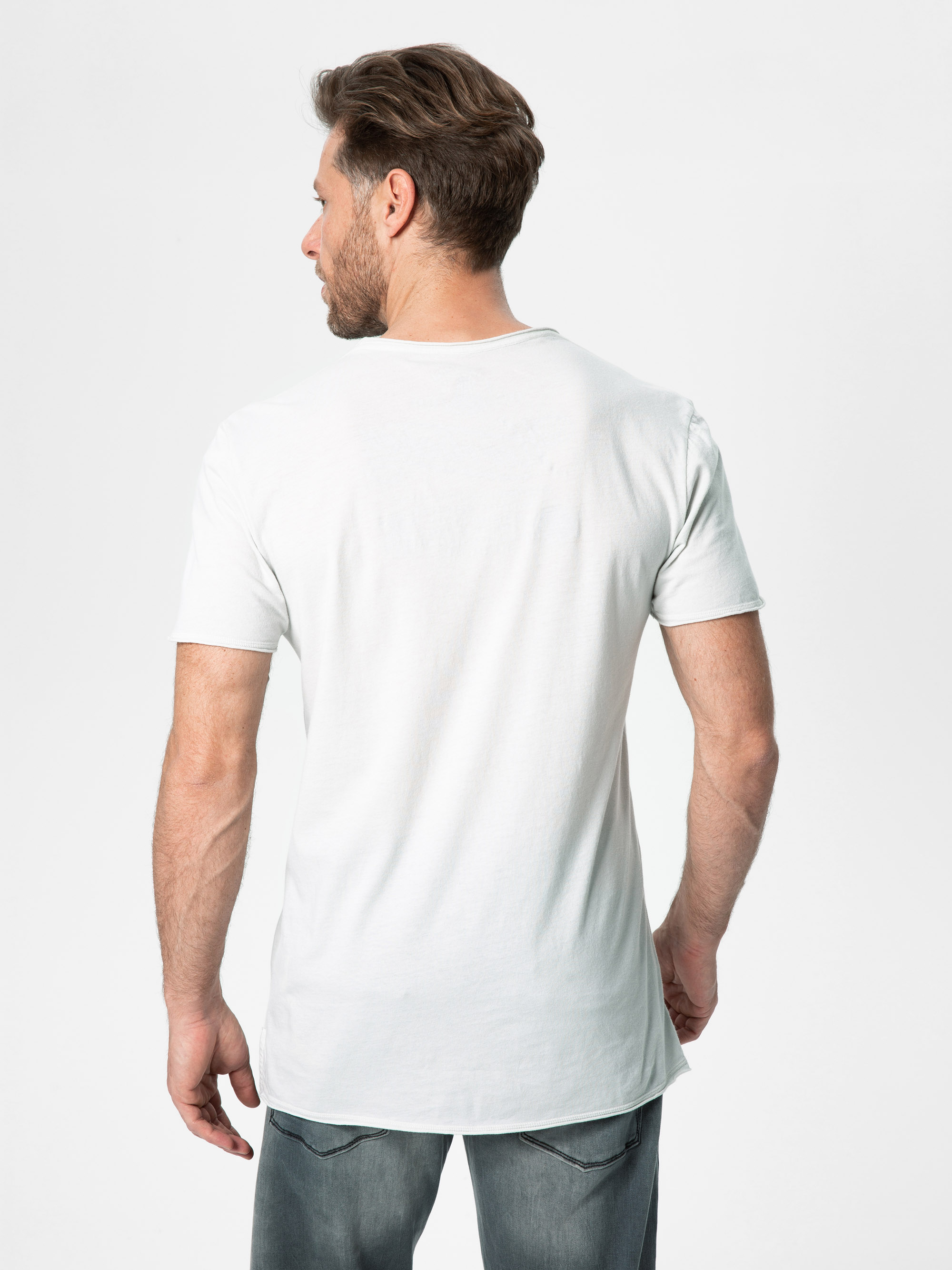 Ein Mann mit kurzen braunen Haaren und Bart steht mit dem Rücken zur Kamera. Er trägt ein schlichtes weißes T-Shirt und verblichene Bluejeans vor einem weißen Hintergrund.