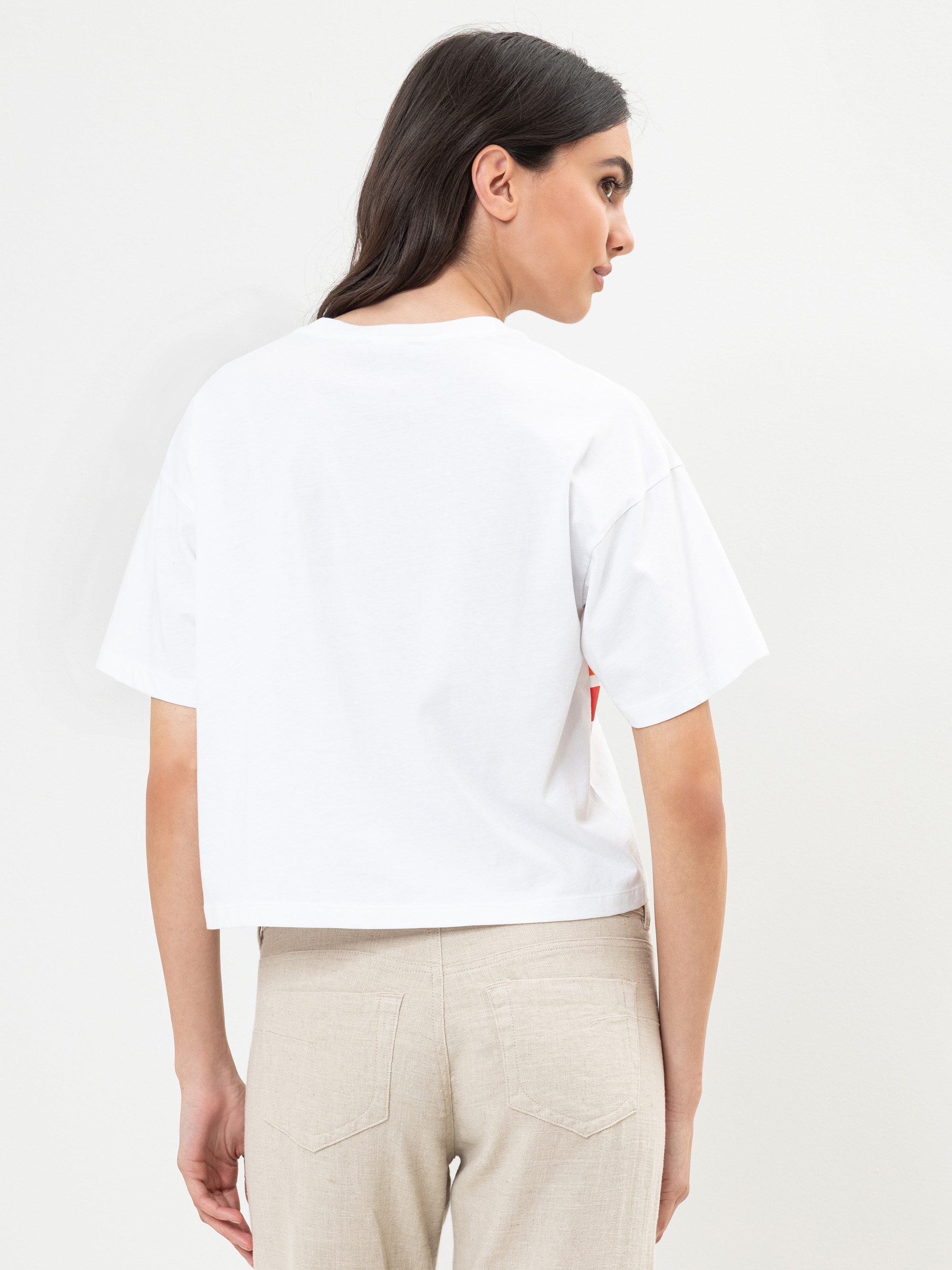 Eine Frau mit langen dunklen Haaren trägt ein lockeres weißes T-Shirt und eine beigefarbene Hose und steht mit dem Rücken zur Kamera vor einem schlichten weißen Hintergrund.