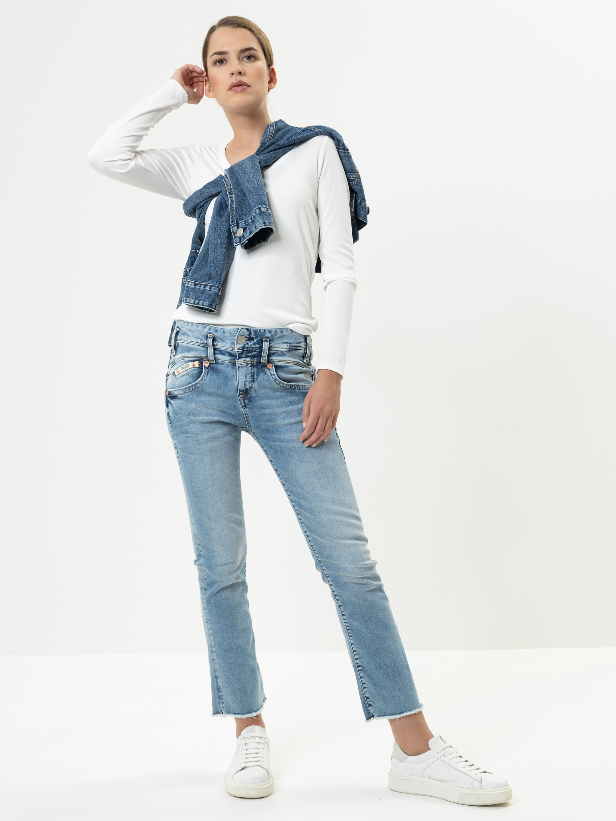 Eine Frau steht selbstbewusst vor einem weißen Hintergrund. Sie trägt ein weißes langärmeliges Hemd, hellblaue Jeans, weiße Turnschuhe und eine Jeansjacke, die sie über die Schultern gelegt hat.