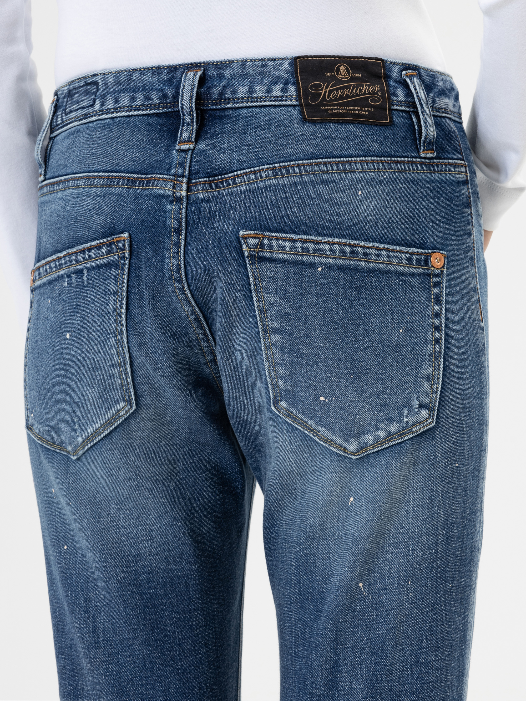 Eine Person, die eine blaue Jeans und ein weißes Hemd trägt, wird von der Taille abwärts gezeigt, wobei der Schwerpunkt auf den Gesäßtaschen und einem braunen Lederaufnäher am Hosenbund liegt.