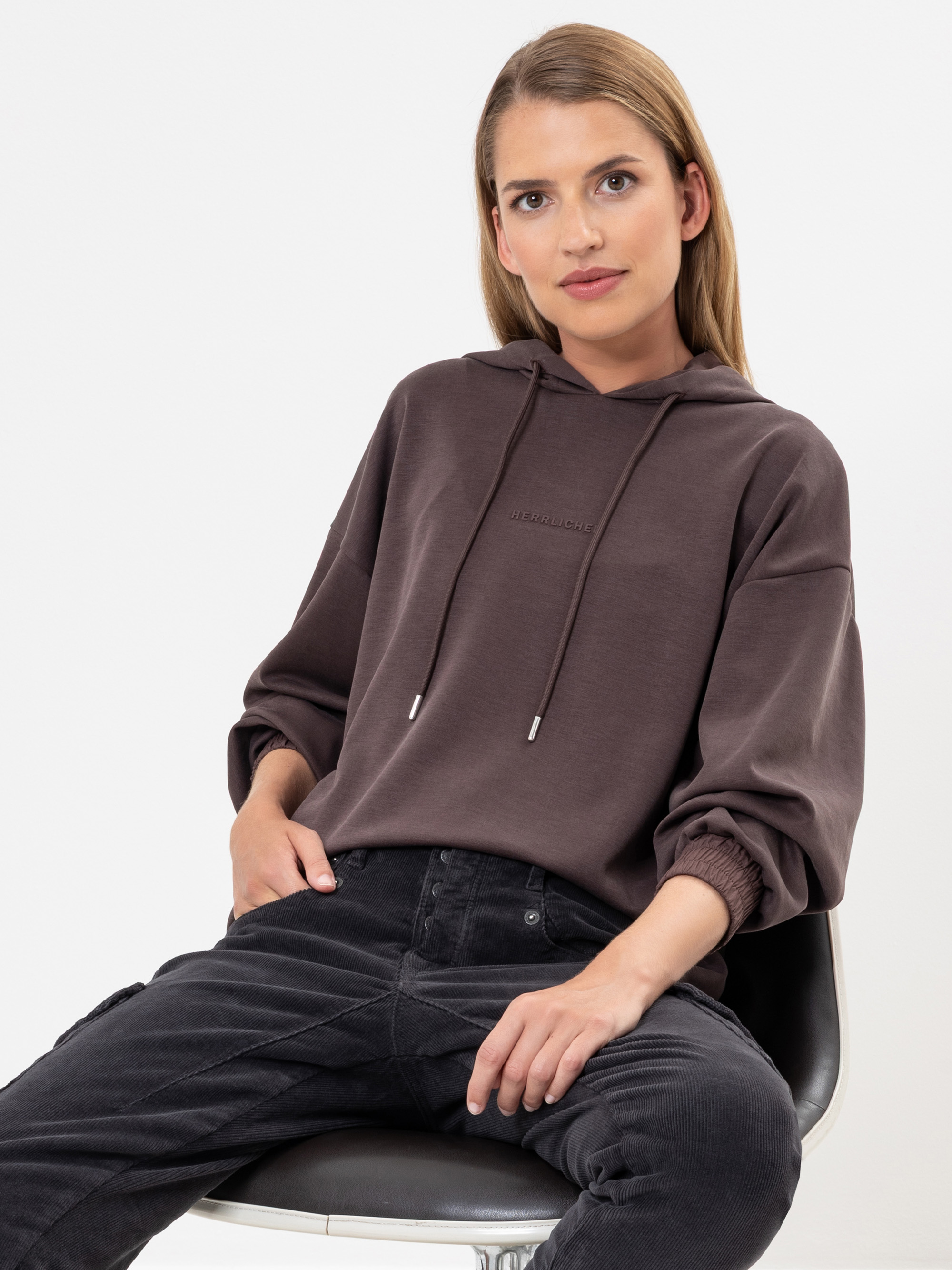 Eine Frau mit glattem blondem Haar sitzt auf einem schwarzen Stuhl, trägt ein braunes Sweatshirt mit Kapuze und dunkle Jeans und blickt in die Kamera vor einem schlichten weißen Hintergrund.