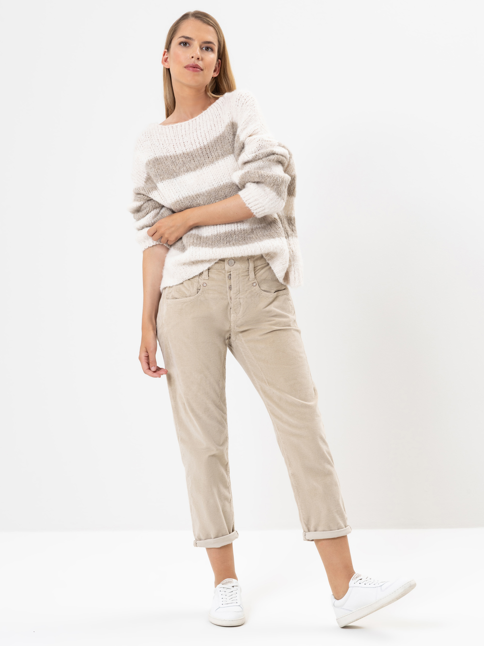Eine Frau steht vor einem schlichten Hintergrund und trägt einen lockeren, gestreiften beigen und weißen Pullover, eine hellbeige Hose mit aufgerollten Bündchen und weiße Turnschuhe. Sie hat glattes Haar und eine entspannte Pose mit einer Hand in der