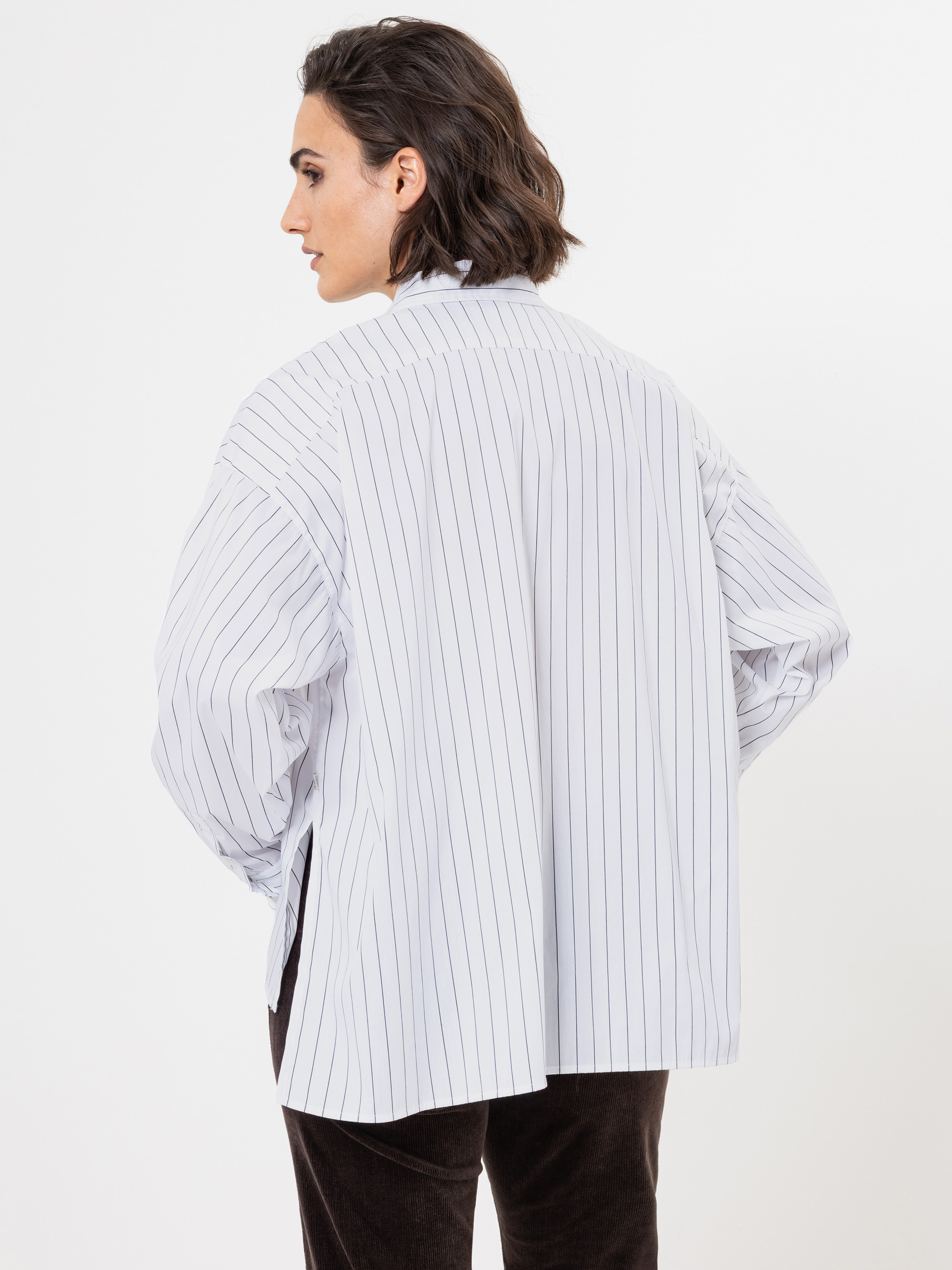 Gezeigt wird eine Person mit kurzen dunklen Haaren von hinten, die ein locker sitzendes weißes Hemd mit dünnen vertikalen schwarzen Streifen und eine dunkle Hose trägt und vor einem schlichten weißen Hintergrund steht.