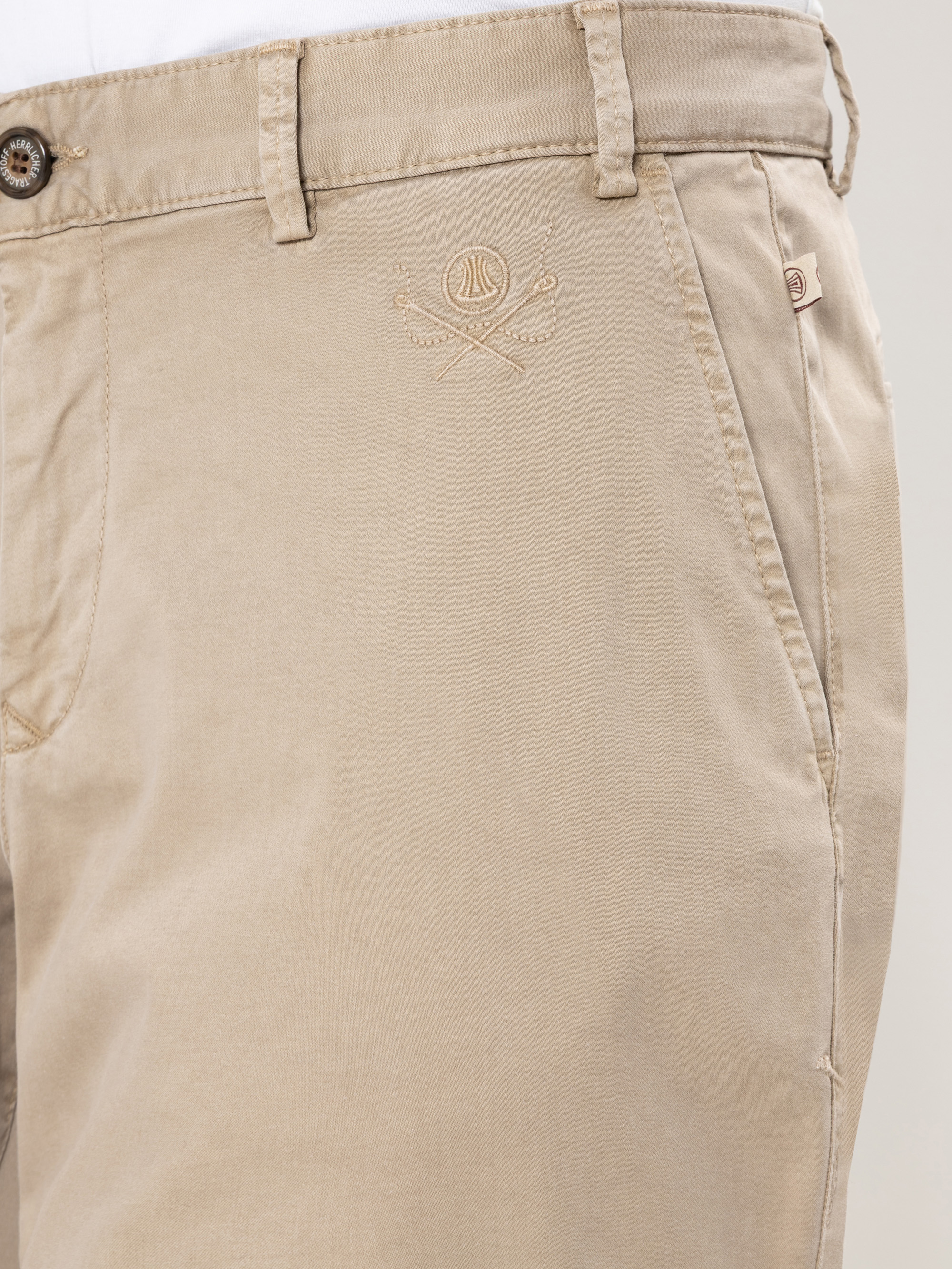 Nahaufnahme einer beigefarbenen khakifarbenen Hose mit Vordertasche, Gürtelschlaufen, Knopf und einem gestickten Logo mit gekreuzten Golfschlägern und einem Golfball auf dem Oberschenkelbereich.