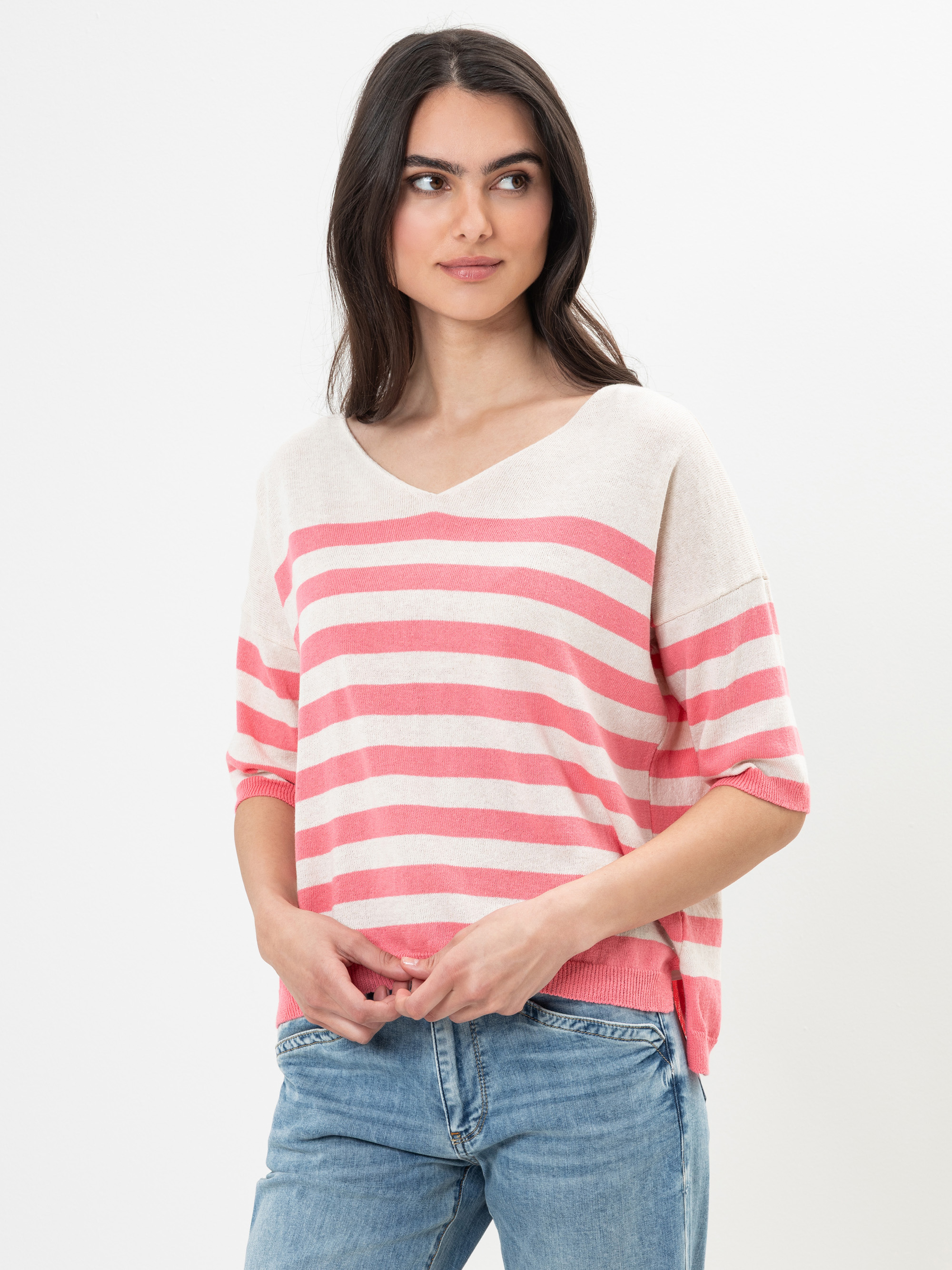 Eine Frau mit langen dunklen Haaren trägt einen lockeren, hellen Pullover mit breiten rosa Querstreifen und blaue Jeans. Sie steht vor einem schlichten weißen Hintergrund und blickt mit neutralem Blick leicht zur Seite.