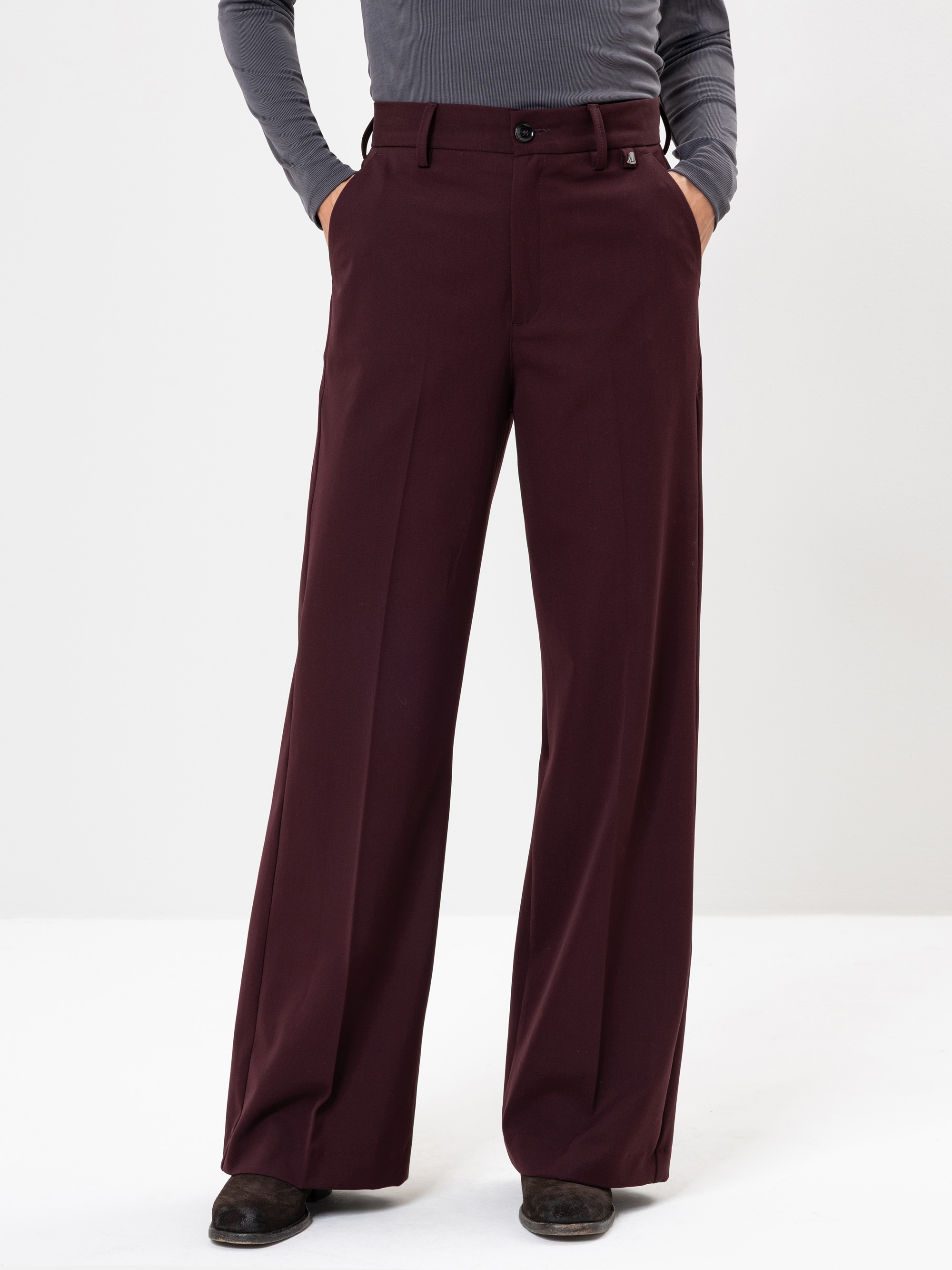 Eine Person, die eine burgunderfarbene Hose mit hoher Taille und weitem Bein trägt, die Hände in den Taschen hat und dazu ein graues, langärmeliges Oberteil und schwarze Schuhe trägt, steht vor einem schlichten weißen Hintergrund.