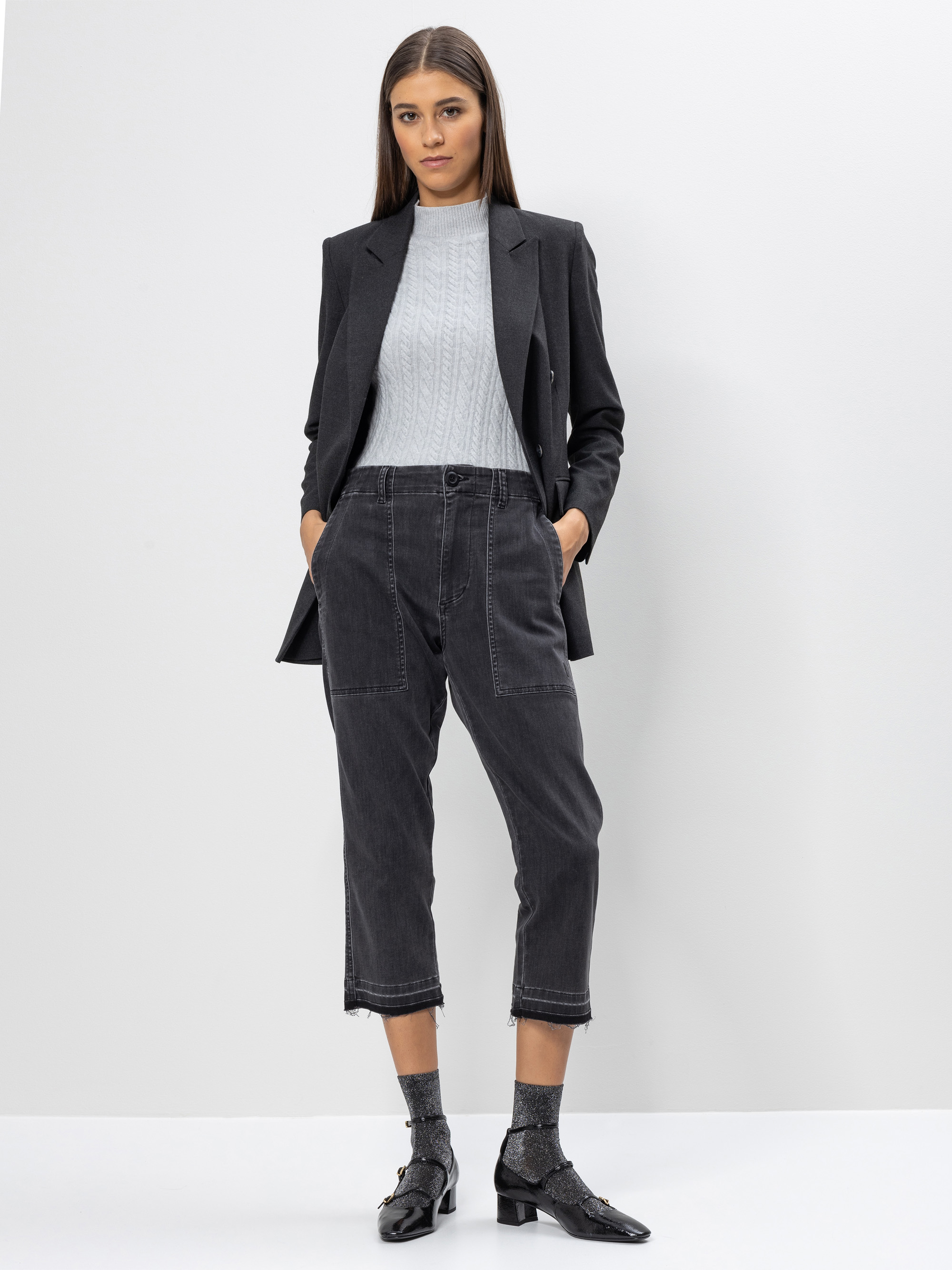 Eine Frau steht vor einem schlichten weißen Hintergrund und trägt einen dunklen Blazer, einen hellgrauen Pullover, eine abgeschnittene schwarze Jeans, glitzernde Socken und schwarze Lackschuhe. Sie hat glattes braunes Haar und ihre Hände stecken in den