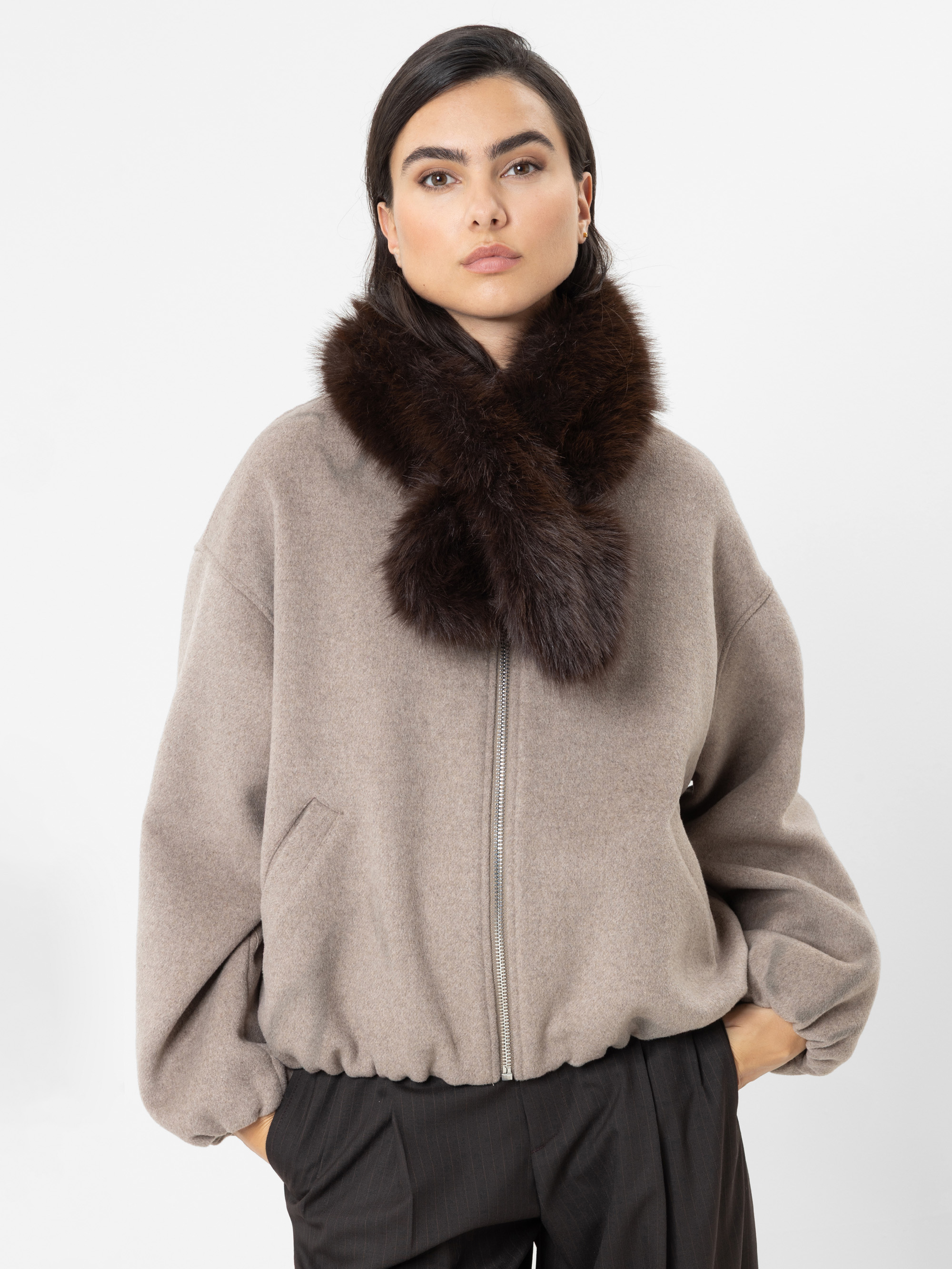 Eine Frau, die eine taupefarbene Jacke mit Reißverschluss, einen dunkelbraunen Pelzschal und eine schwarze Hose trägt, steht vor einem schlichten weißen Hintergrund und blickt mit neutralem Gesichtsausdruck direkt in die Kamera.