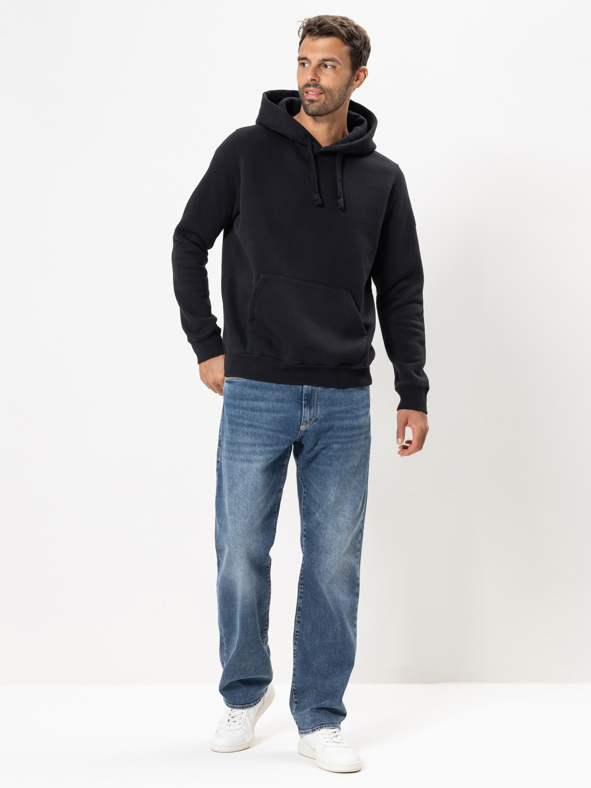 Ein Mann steht vor einem schlichten weißen Hintergrund und trägt einen schwarzen Kapuzenpullover, blaue Jeans und weiße Turnschuhe. Er hat kurzes dunkles Haar, einen gestutzten Bart und blickt mit entspannter Miene leicht nach links.