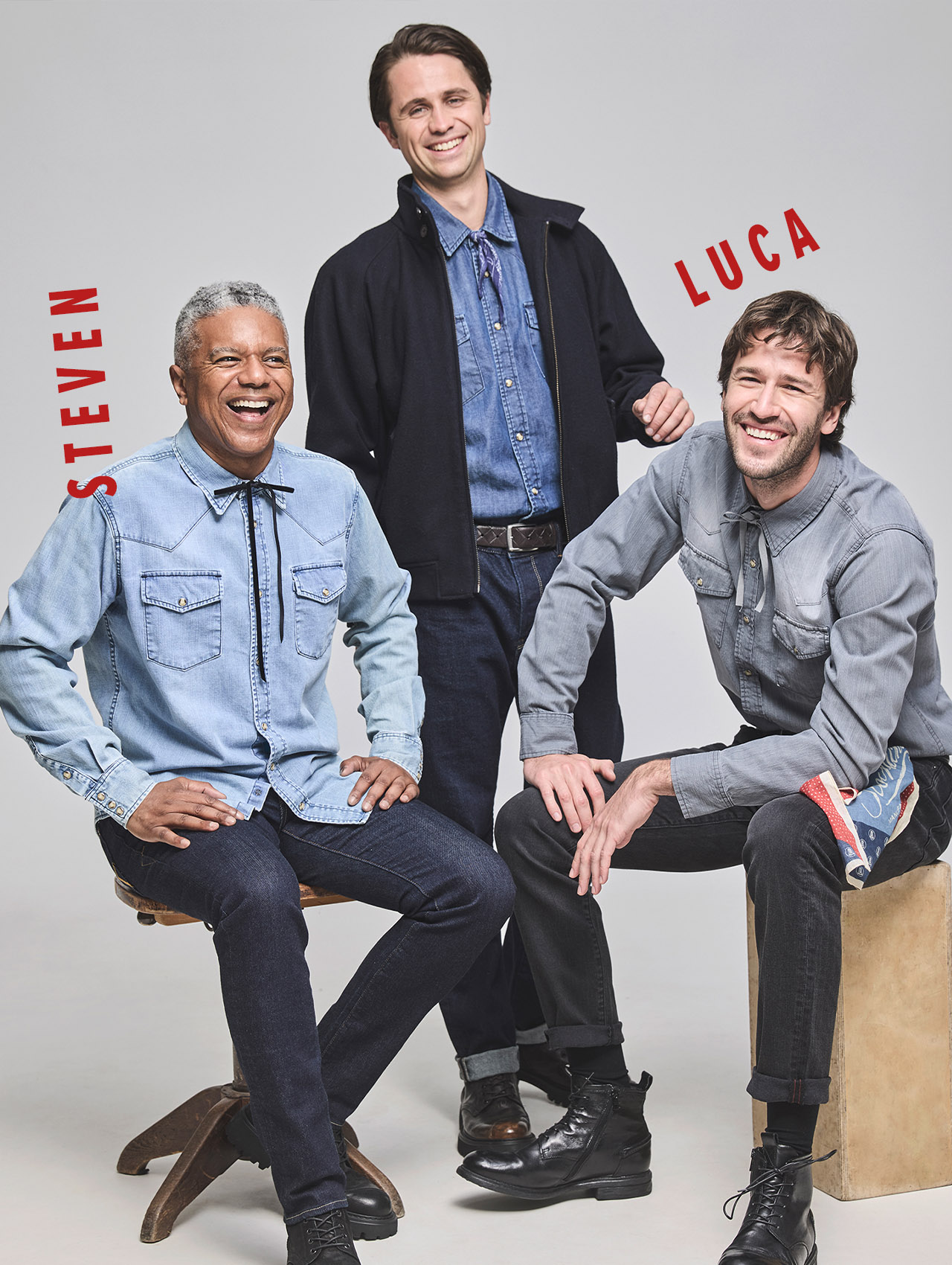 Drei Männer in Jeans-Outfits posieren und lächeln vor einem schlichten Hintergrund. Die beiden Sitzenden sitzen auf Hockern oder Kisten, der Stehende steht hinter ihnen. Der rote Text auf dem Bild lautet STEVEN und LUCA. Alle sind leger gekleidet.