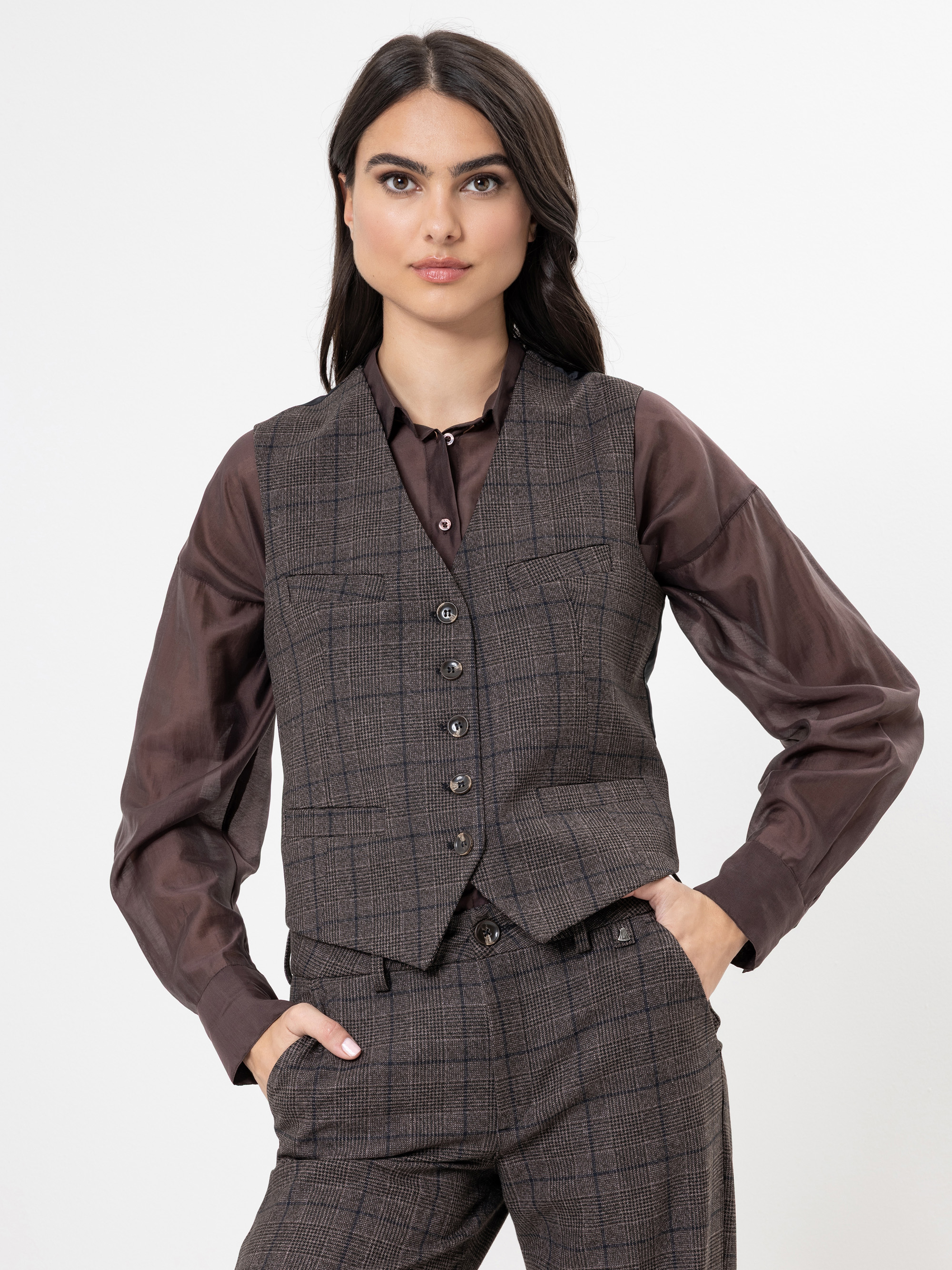 Eine Frau mit langen dunklen Haaren trägt eine braun karierte Weste und eine dazu passende Hose über einer dunkelbraunen Bluse. Sie steht vor einem schlichten weißen Hintergrund, eine Hand in der Tasche, und schaut selbstbewusst in die Kamera.