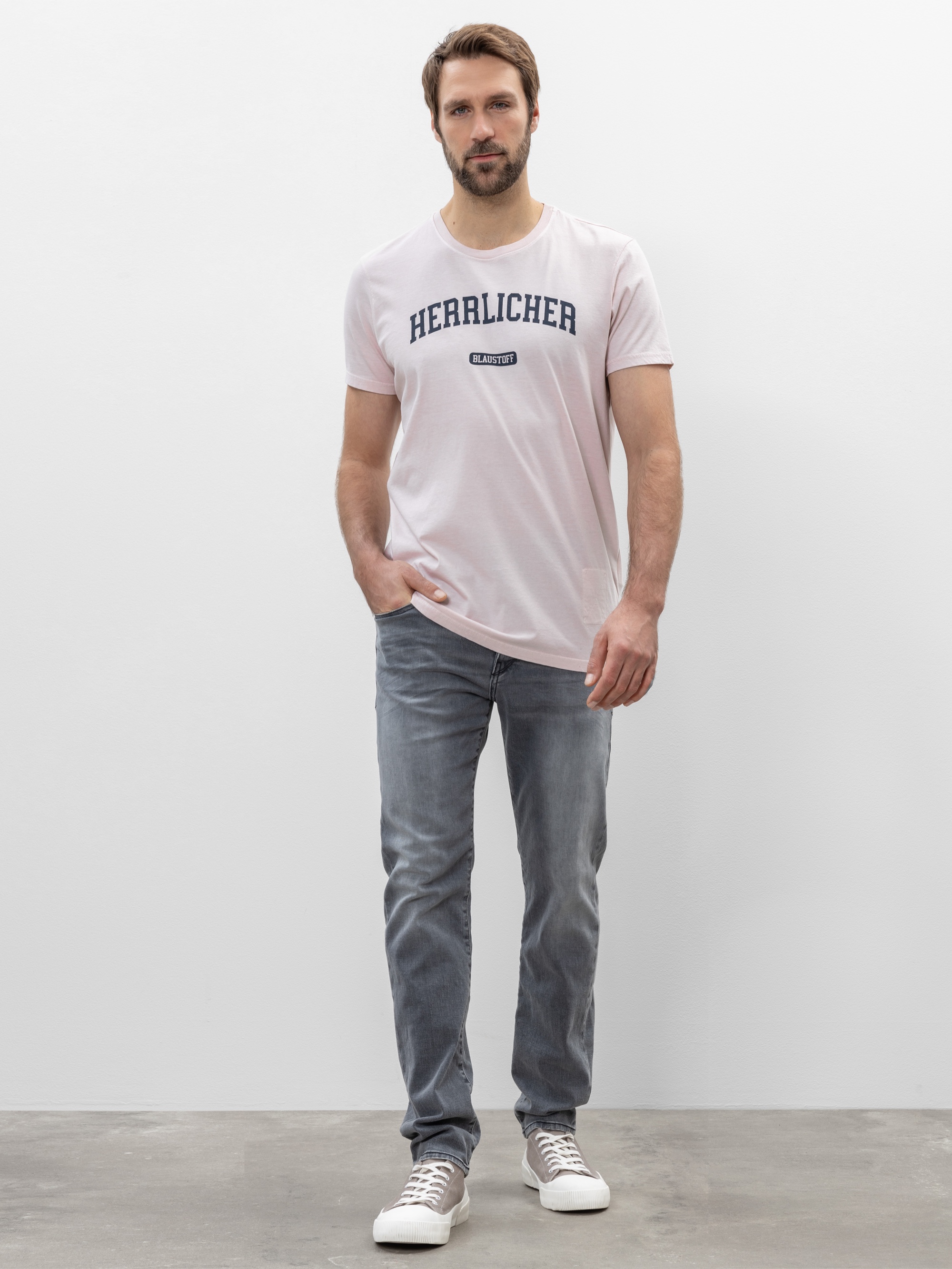 Ein Mann mit kurzen braunen Haaren und einem Bart trägt ein hellrosa T-Shirt mit dem Aufdruck HERALICHER, graue Jeans und weiße Turnschuhe. Er steht vor einer schlichten weißen Wand auf einem grauen Boden.