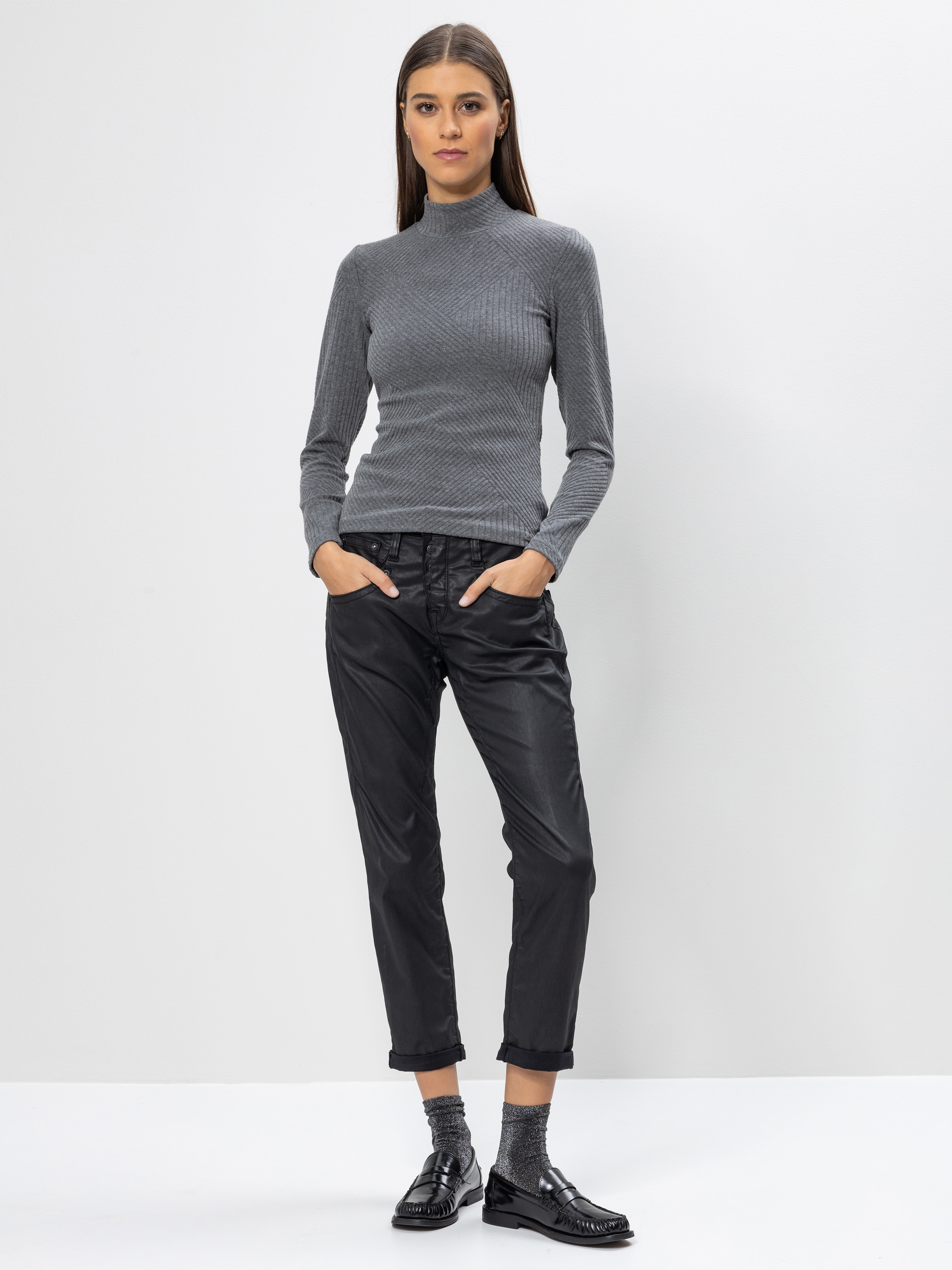 Eine Frau steht vor einem schlichten weißen Hintergrund und trägt einen grauen Rollkragenpullover, eine schwarze Hose mit Bündchen und schwarze, glänzende Schuhe mit Socken. Ihre Hände stecken in den Taschen, und sie hat glattes, dunkles Haar.