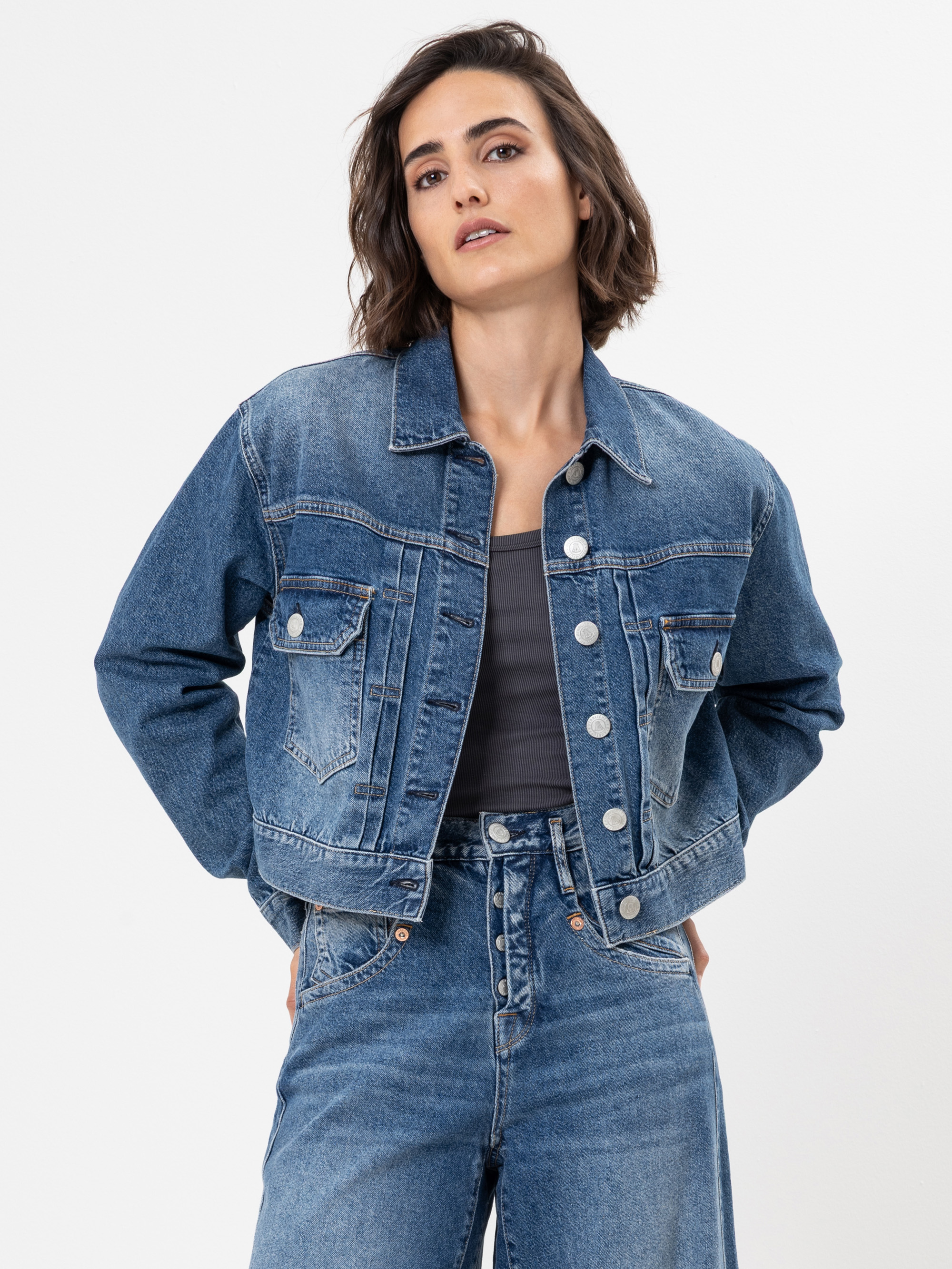 Eine Frau mit kurzen braunen Haaren, die eine blaue Jeansjacke und hochgekrempelte Jeans trägt, steht vor einem weißen Hintergrund und blickt selbstbewusst in die Kamera, die Hände in die Hüften gestemmt.