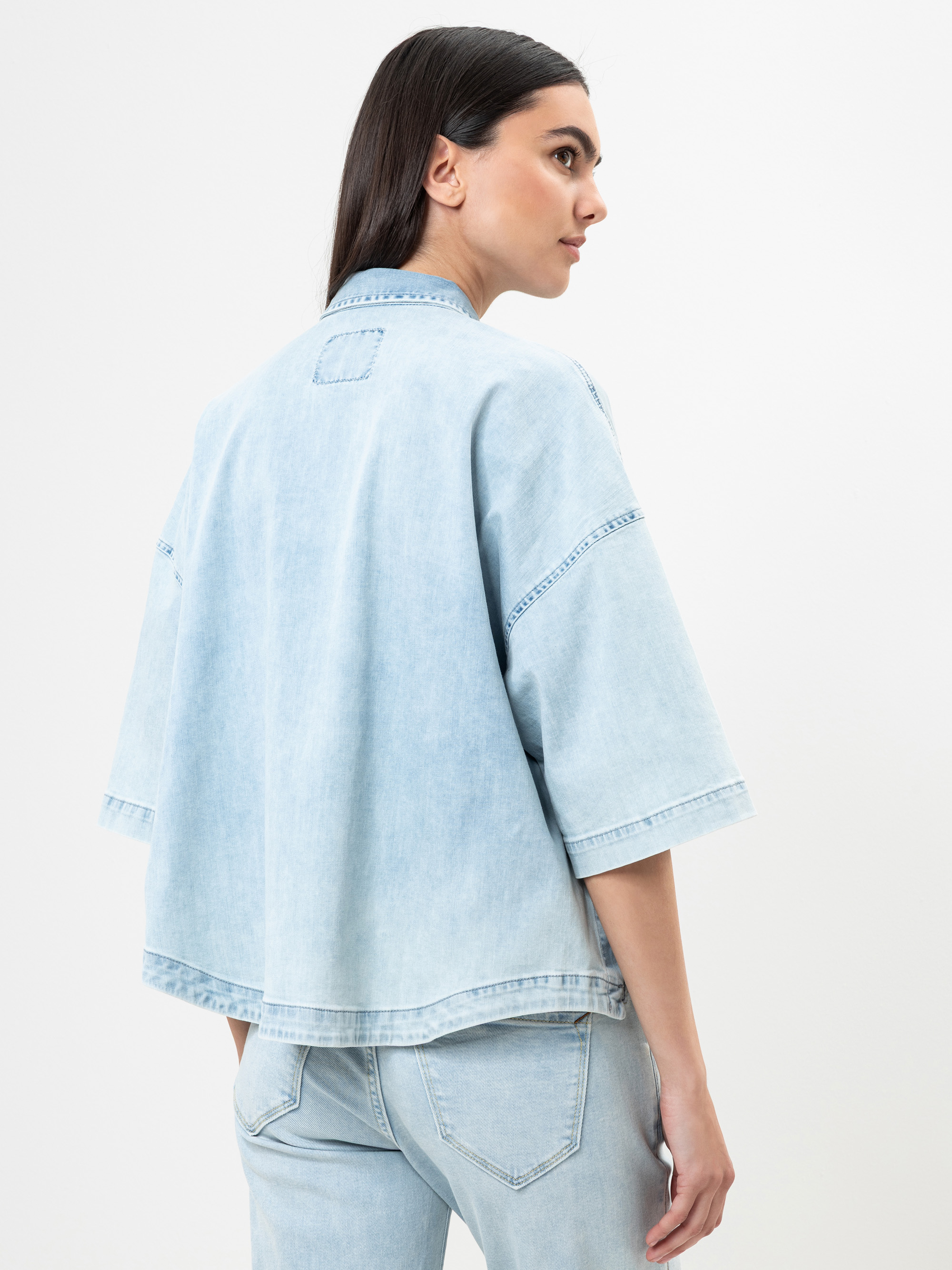 Eine Frau mit langen dunklen Haaren wird von hinten gezeigt. Sie trägt eine hellblaue Jeansjacke mit weiten Ärmeln und dazu passende hellblaue Jeans vor einem schlichten weißen Hintergrund.