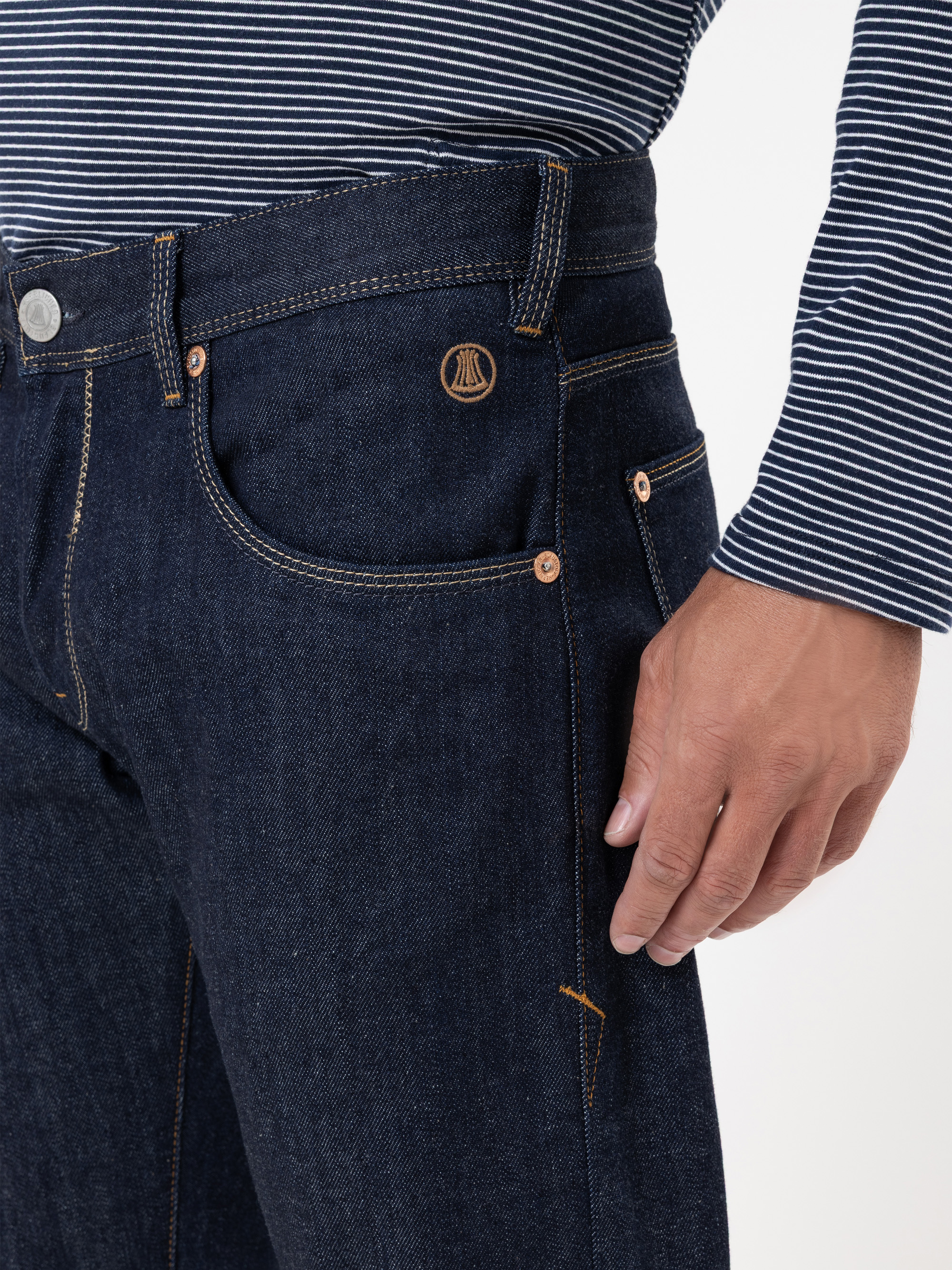 Eine Person, die eine dunkelblaue Jeans und ein marineblau-weiß gestreiftes, langärmeliges Hemd trägt, wobei ihre Hand in der Nähe der Jeanstasche ruht.