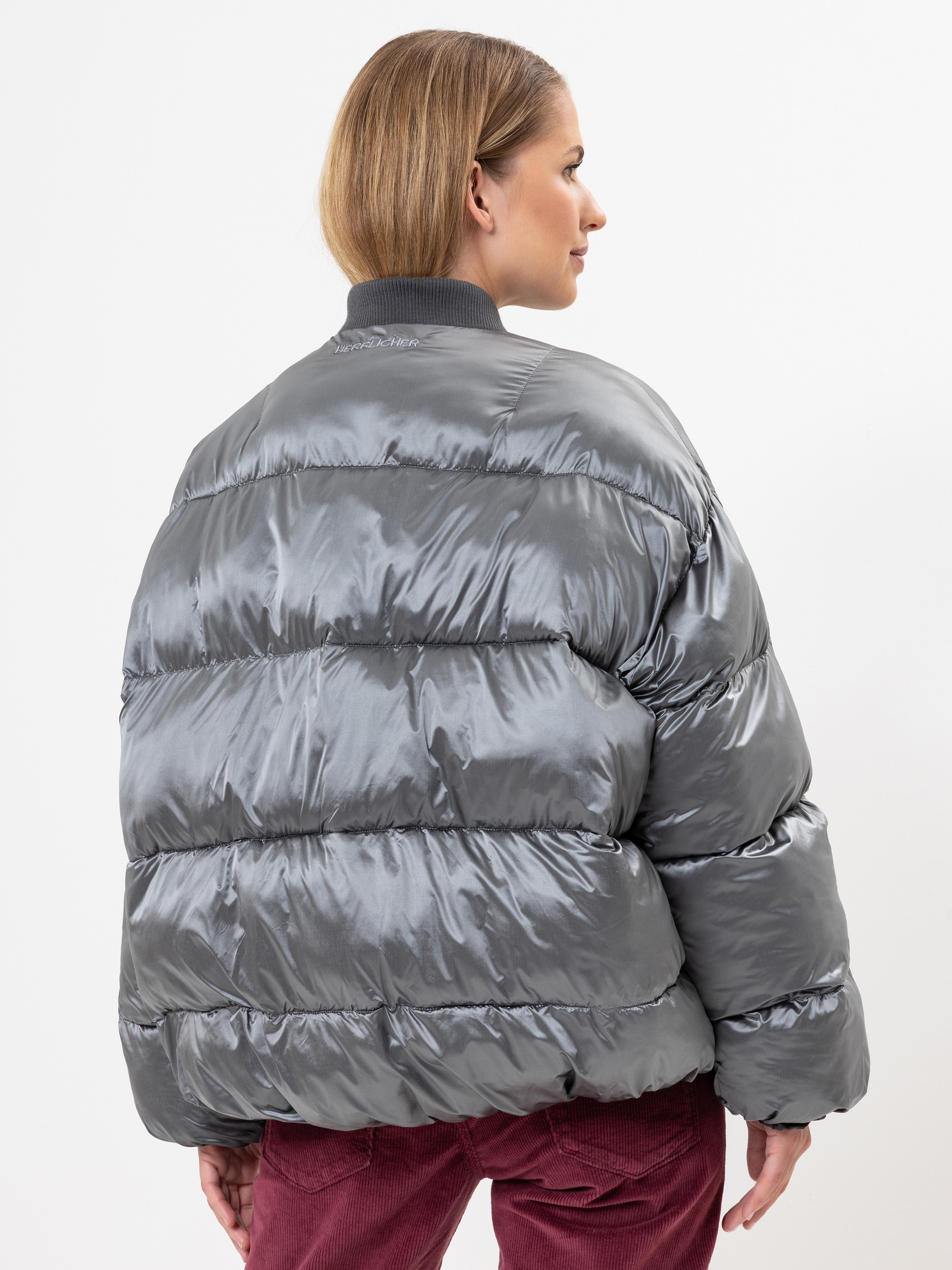 Eine Person mit hellbraunem Haar, von hinten gesehen, trägt eine glänzende graue Pufferjacke und eine burgunderfarbene Hose. Der Hintergrund ist einfarbig weiß.