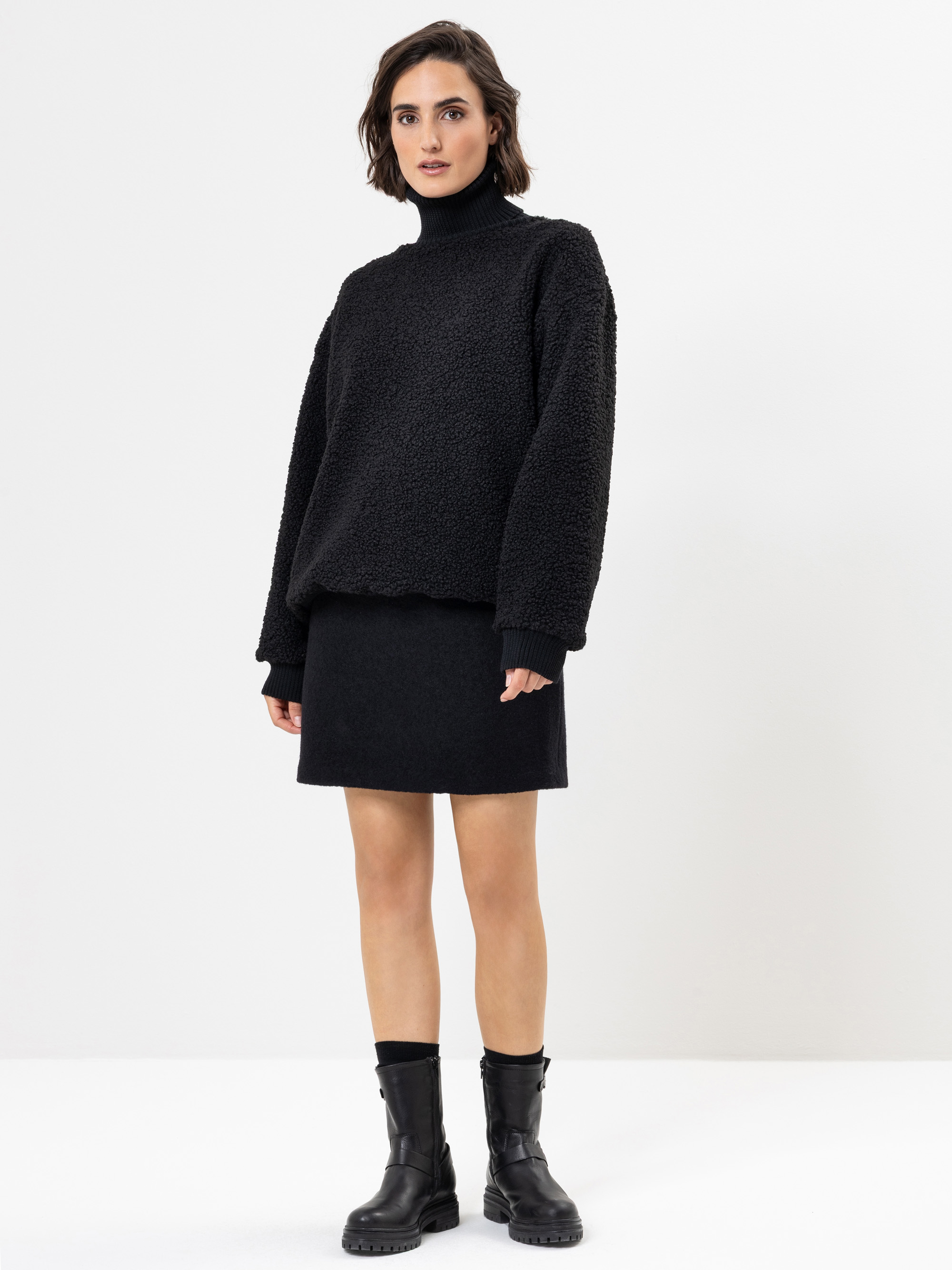 Eine Frau steht vor einem weißen Hintergrund und trägt einen schwarzen Rollkragenpullover, einen schwarzen Rock und schwarze Ankle Boots. Sie hat kurzes braunes Haar und einen neutralen Gesichtsausdruck.