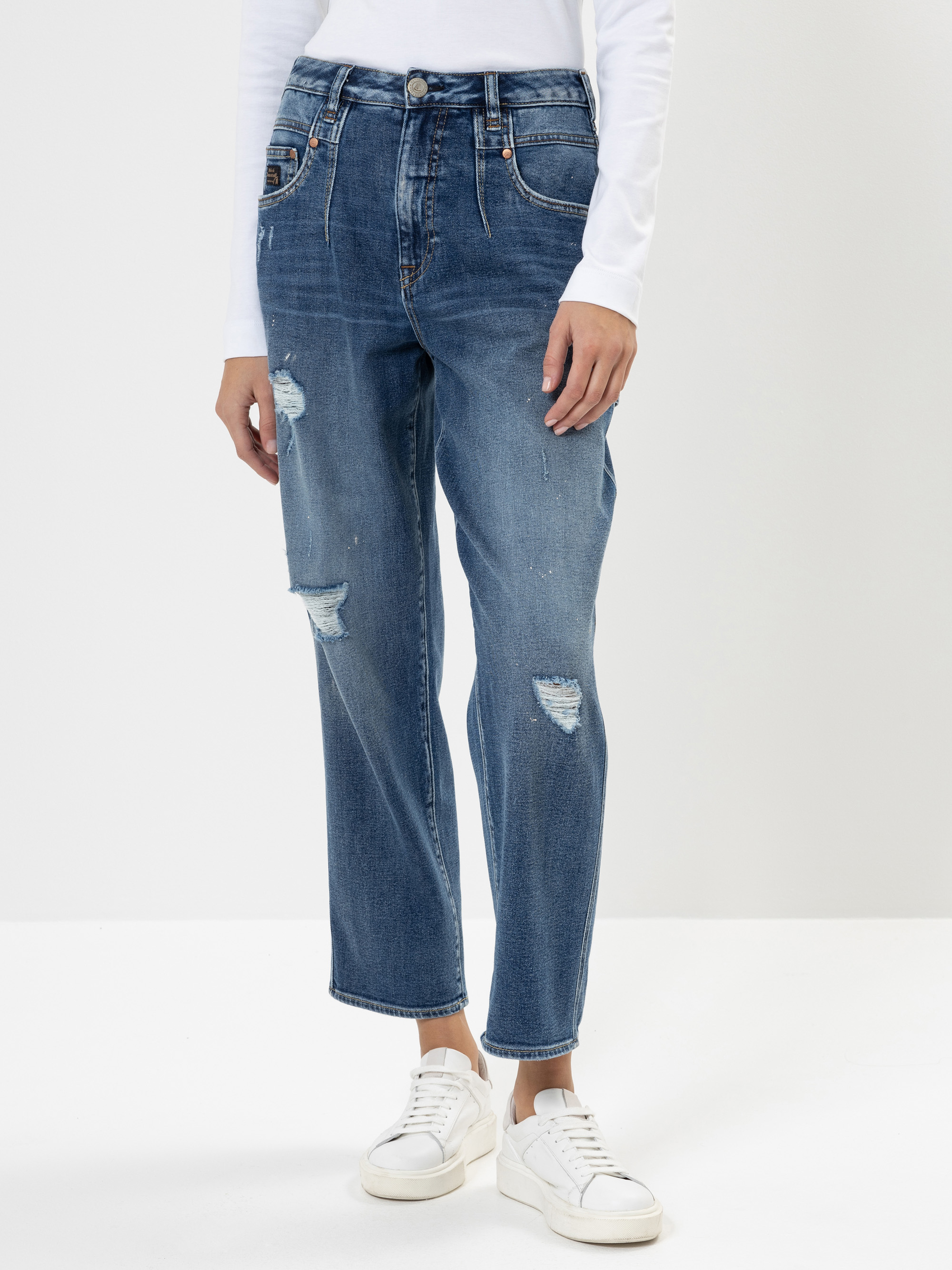 Eine Person, die blaue, zerrissene Jeans mit geradem Bein, ein weißes langärmeliges Oberteil und weiße Turnschuhe trägt, steht vor einem schlichten weißen Hintergrund. Das Bild ist an den Schultern abgeschnitten.