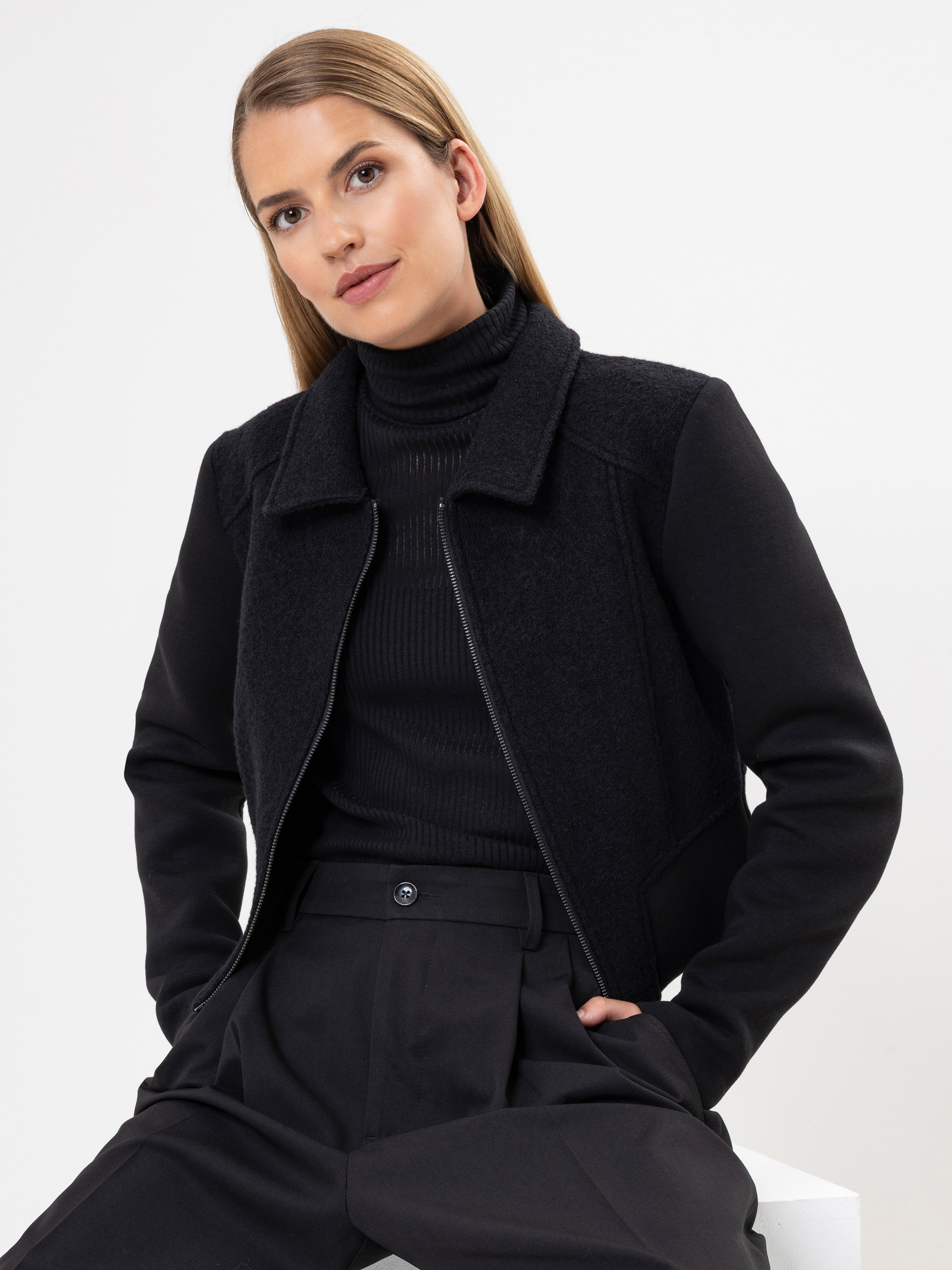 Eine Frau mit glattem, hellbraunem Haar trägt eine schwarze Jacke mit Reißverschluss über einem schwarzen Rollkragenpullover und eine schwarze Hose mit hoher Taille. Sie sitzt leicht angewinkelt, die Hände in den Taschen, vor einem schlichten weißen