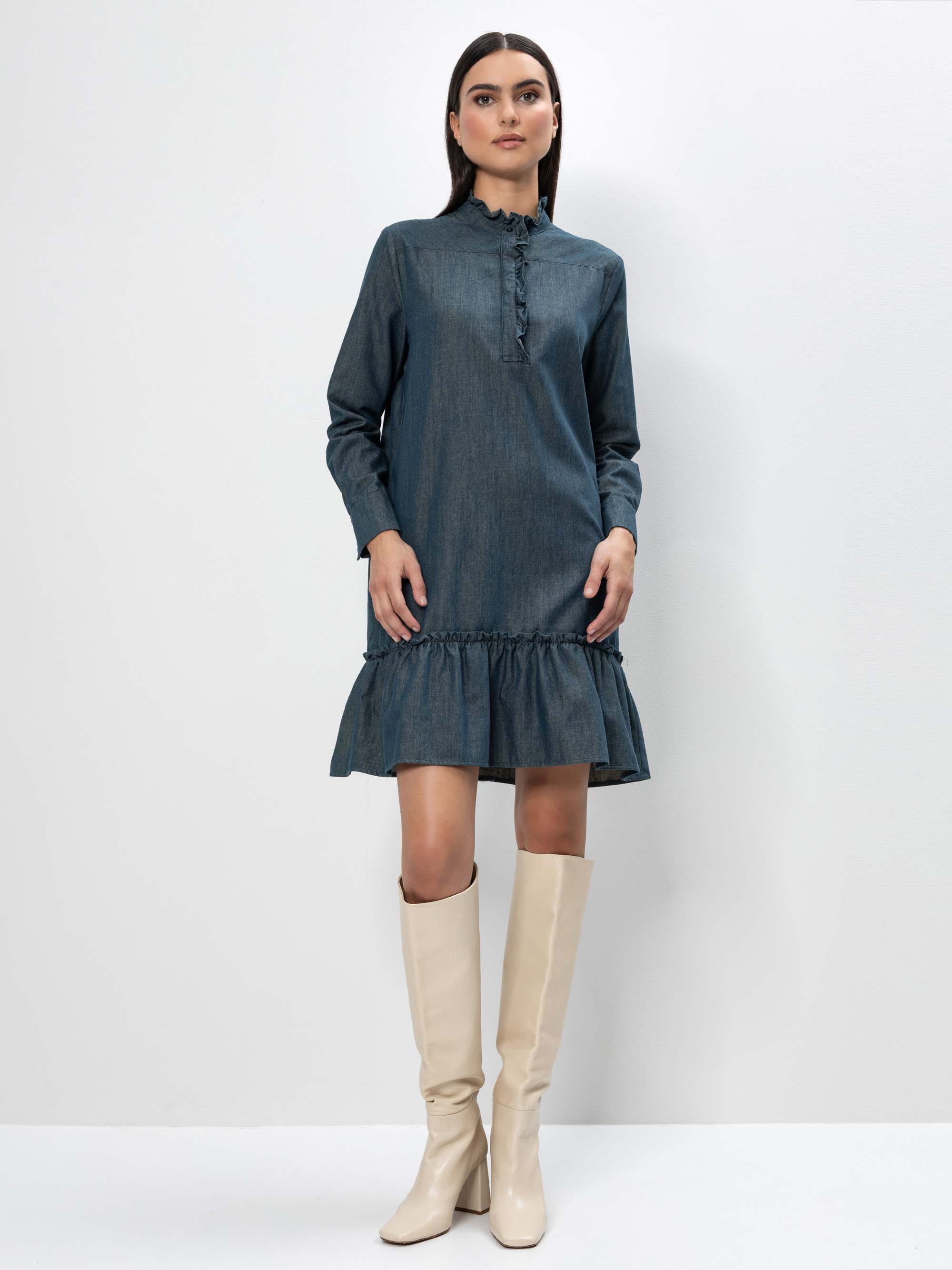 Eine Frau steht vor einem weißen Hintergrund und trägt ein langärmeliges, blaues Jeanskleid mit Rüschendetails und cremefarbene kniehohe Stiefel. Sie hat glattes dunkles Haar und einen neutralen Gesichtsausdruck.