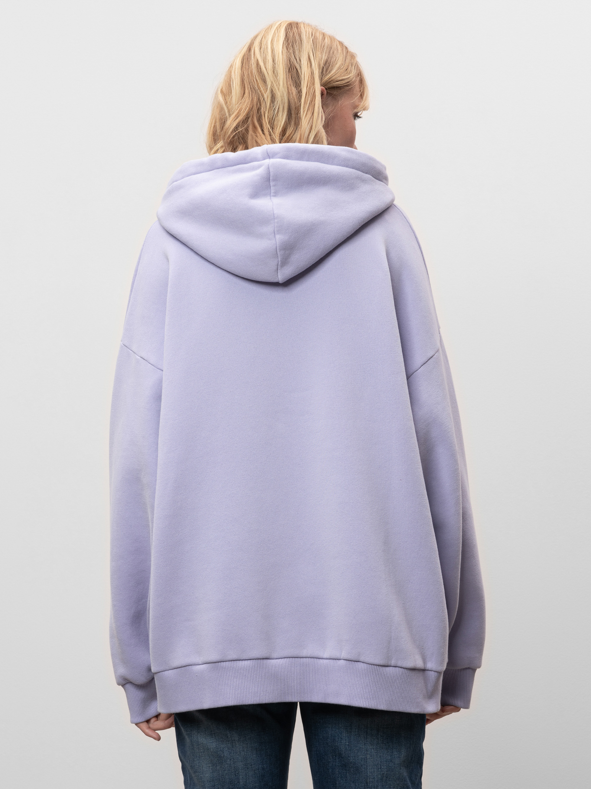 Eine Person mit schulterlangen blonden Haaren wird von hinten gezeigt. Sie trägt einen locker sitzenden, hellvioletten Kapuzenpullover und blaue Jeans vor einem schlichten hellgrauen Hintergrund.