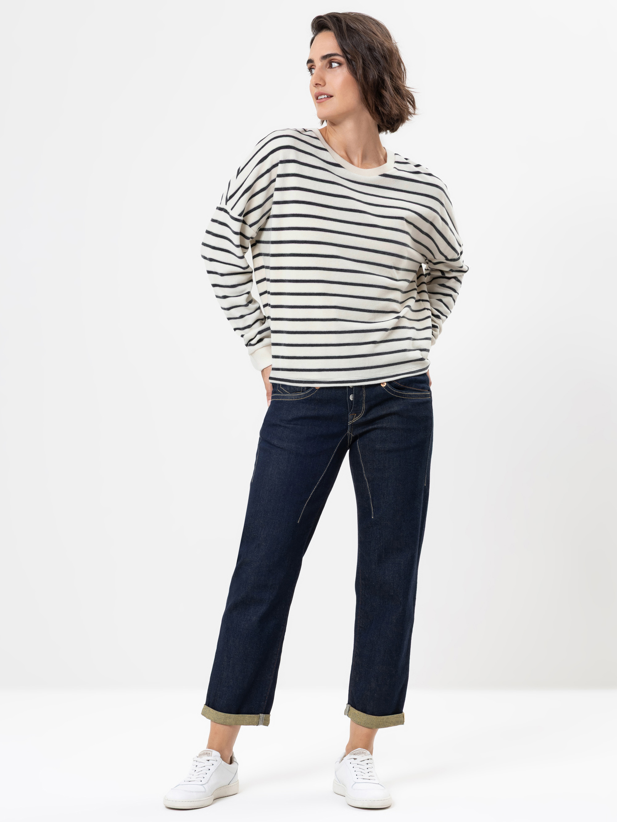 Eine Person, die ein cremefarbenes und schwarz gestreiftes langärmeliges Hemd, dunkle Jeans mit Bündchen und weiße Turnschuhe trägt, steht vor einem schlichten, hellen Hintergrund und blickt mit den Händen in den Taschen zur Seite.