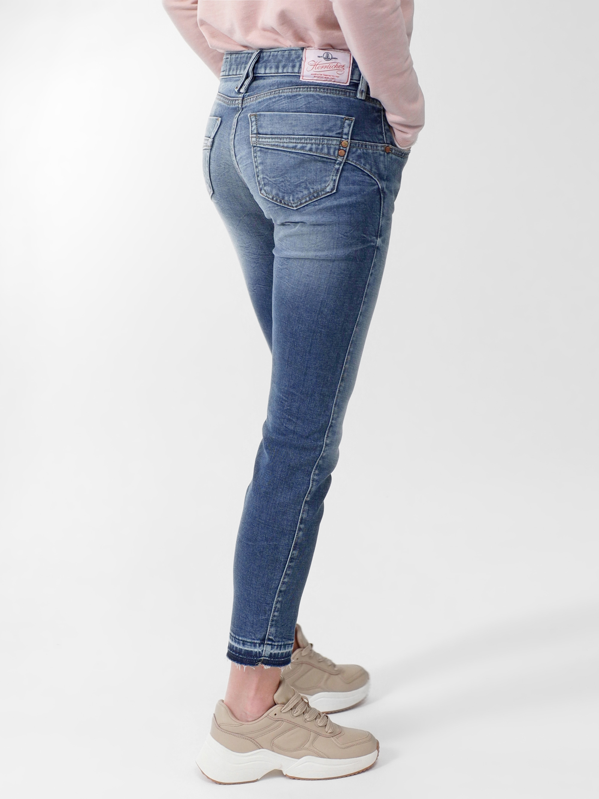 Eine Person in blauen Skinny-Jeans, einem hellrosa Sweatshirt und beigen Turnschuhen steht vor einem schlichten weißen Hintergrund, von hinten und leicht zur Seite gezeigt.